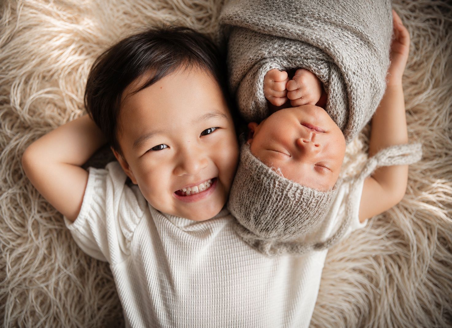 Smiling toddler lying next to sleeping newborn wrapped in gray knit blanket on fluffy white rug.