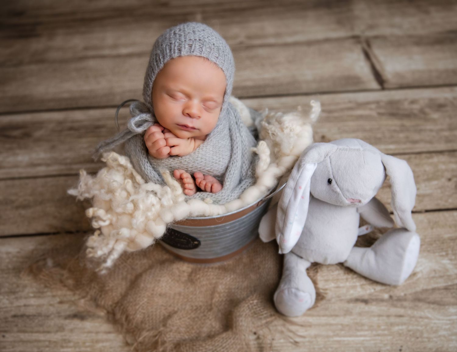 Sleeping newborn baby in gray knit bonnet nestled in metal bucket with white bunny stuffed animal on wood floor.