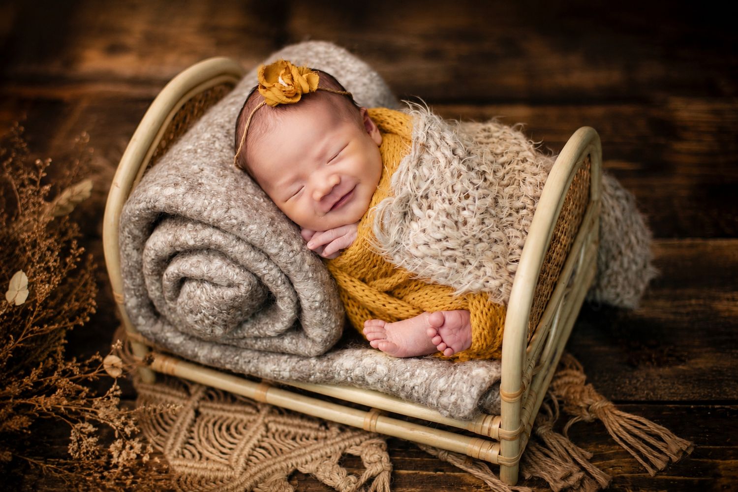 Smiling newborn baby wrapped in gray knit blanket wearing mustard yellow outfit in tiny rattan bed.