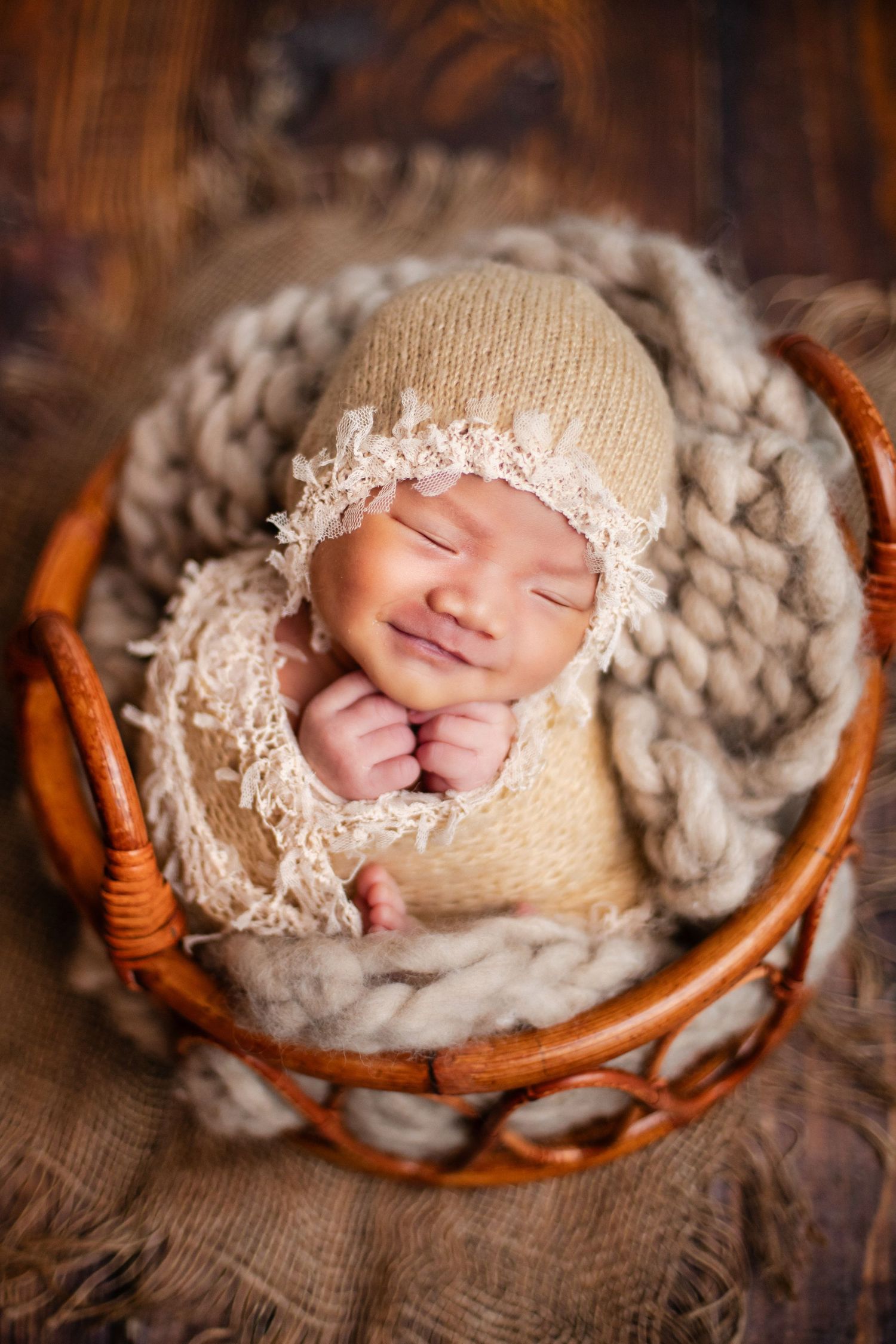 Smiling newborn baby in beige knit outfit and bonnet sleeping in a wicker basket with chunky knit blanket.