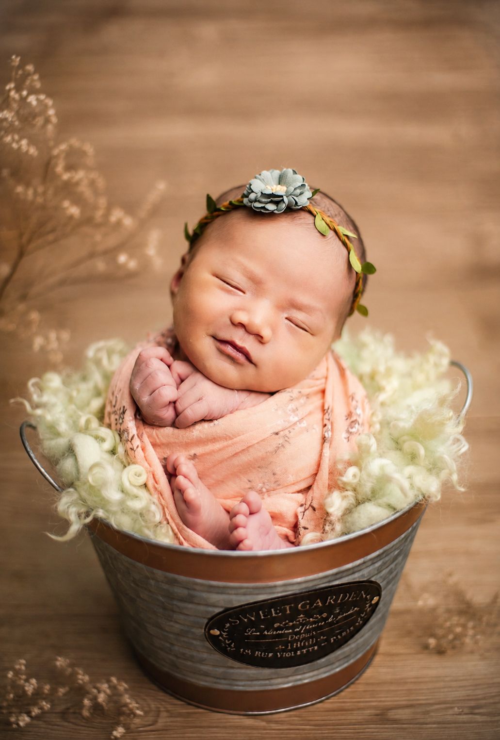 Smiling newborn baby wrapped in peach swaddle with floral headband posed in a rustic metal bucket with white wool.