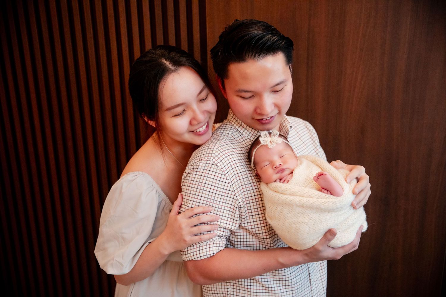 Happy Asian couple smiling and holding their swaddled newborn baby against a wooden panel background.