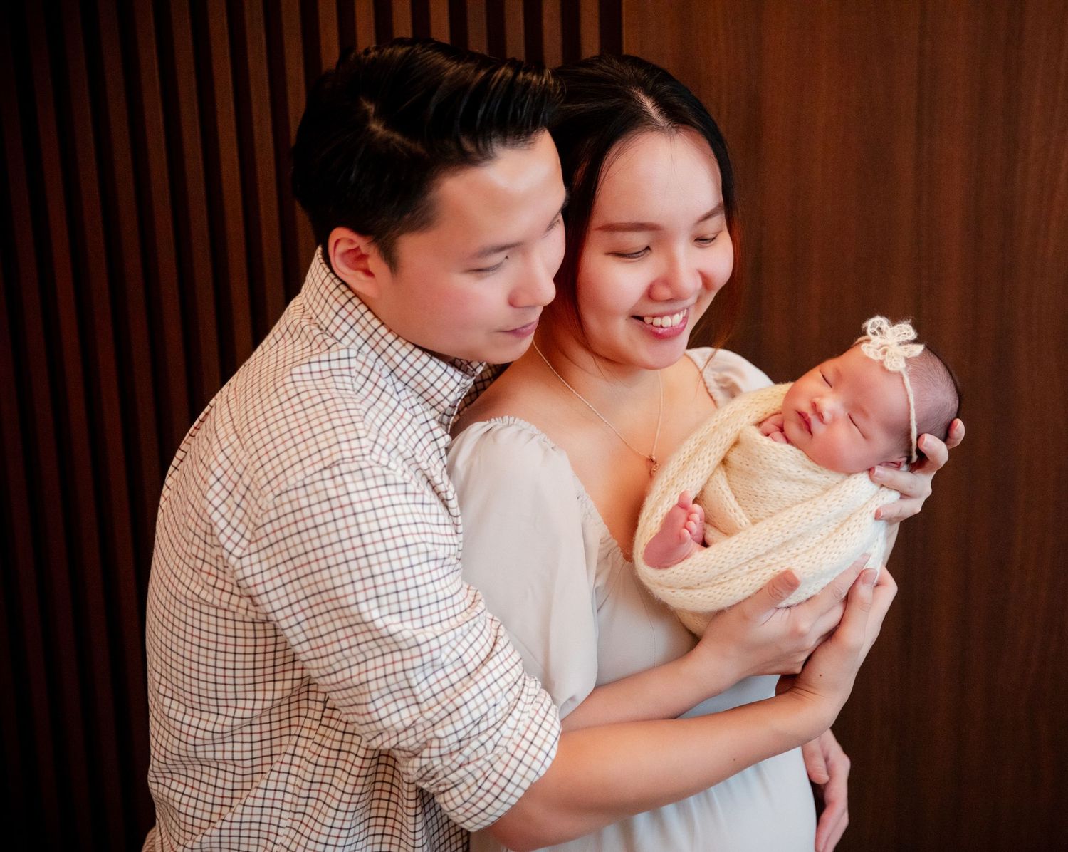 Happy Asian couple smiling and holding their newborn baby wrapped in cream swaddle against wooden backdrop.
