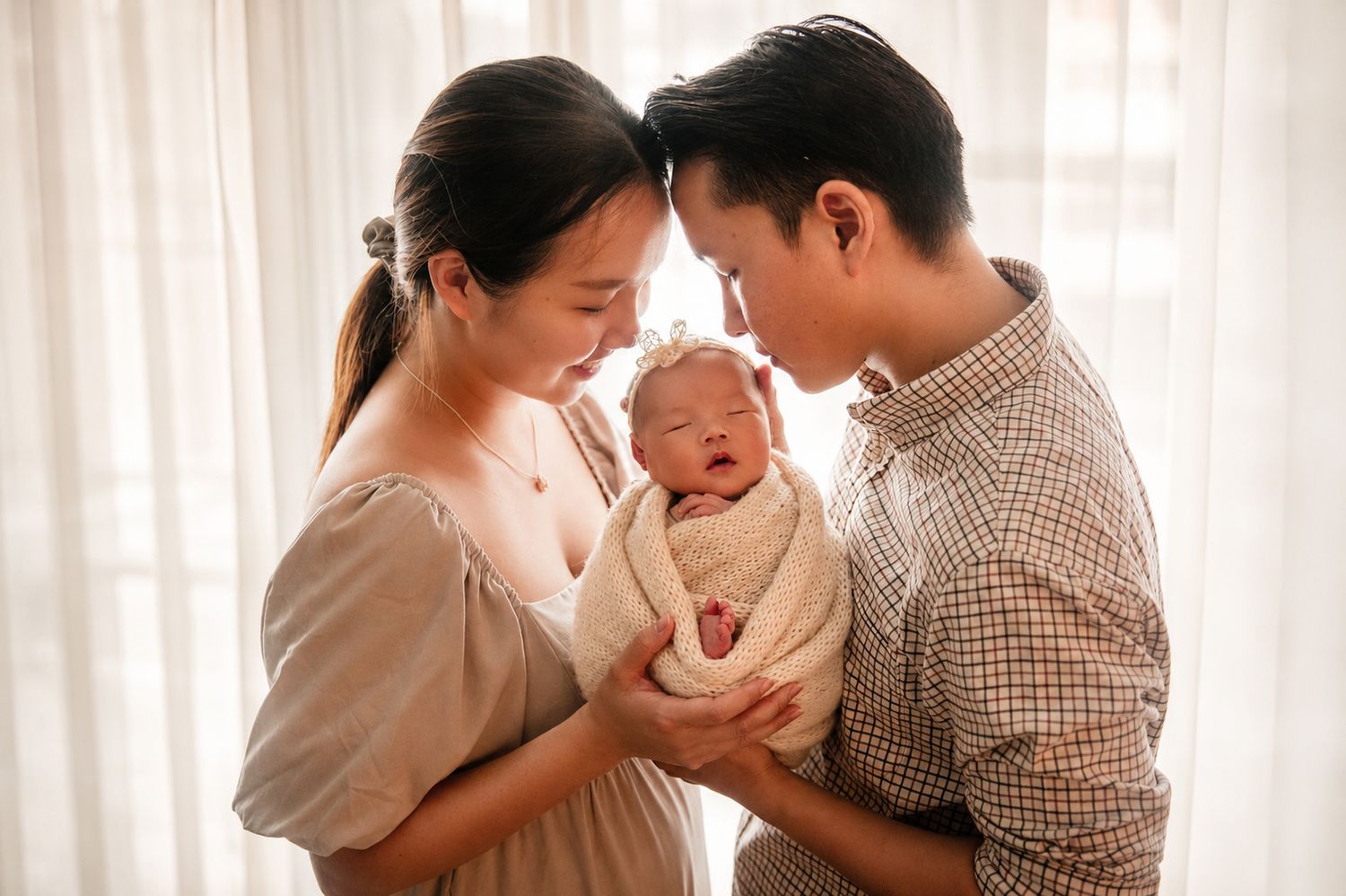 A couple tenderly holds their newborn baby, touching foreheads in a bright, softly lit room.
