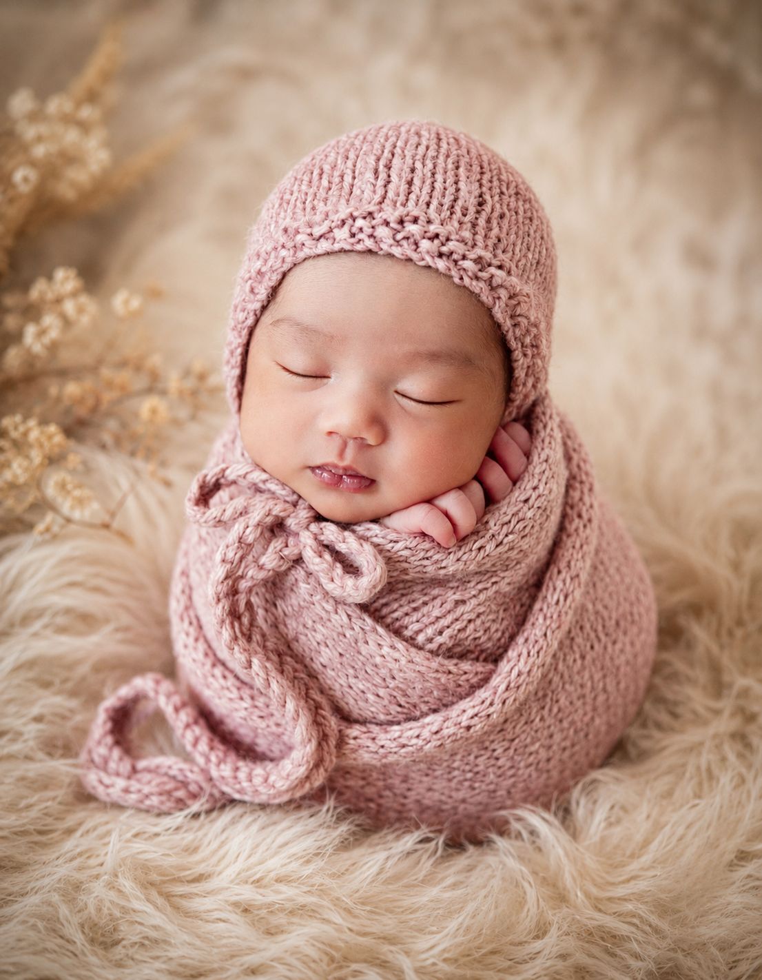 Sleeping newborn baby wrapped in a soft pink knit blanket and matching bonnet hat on a fluffy cream fur rug.
