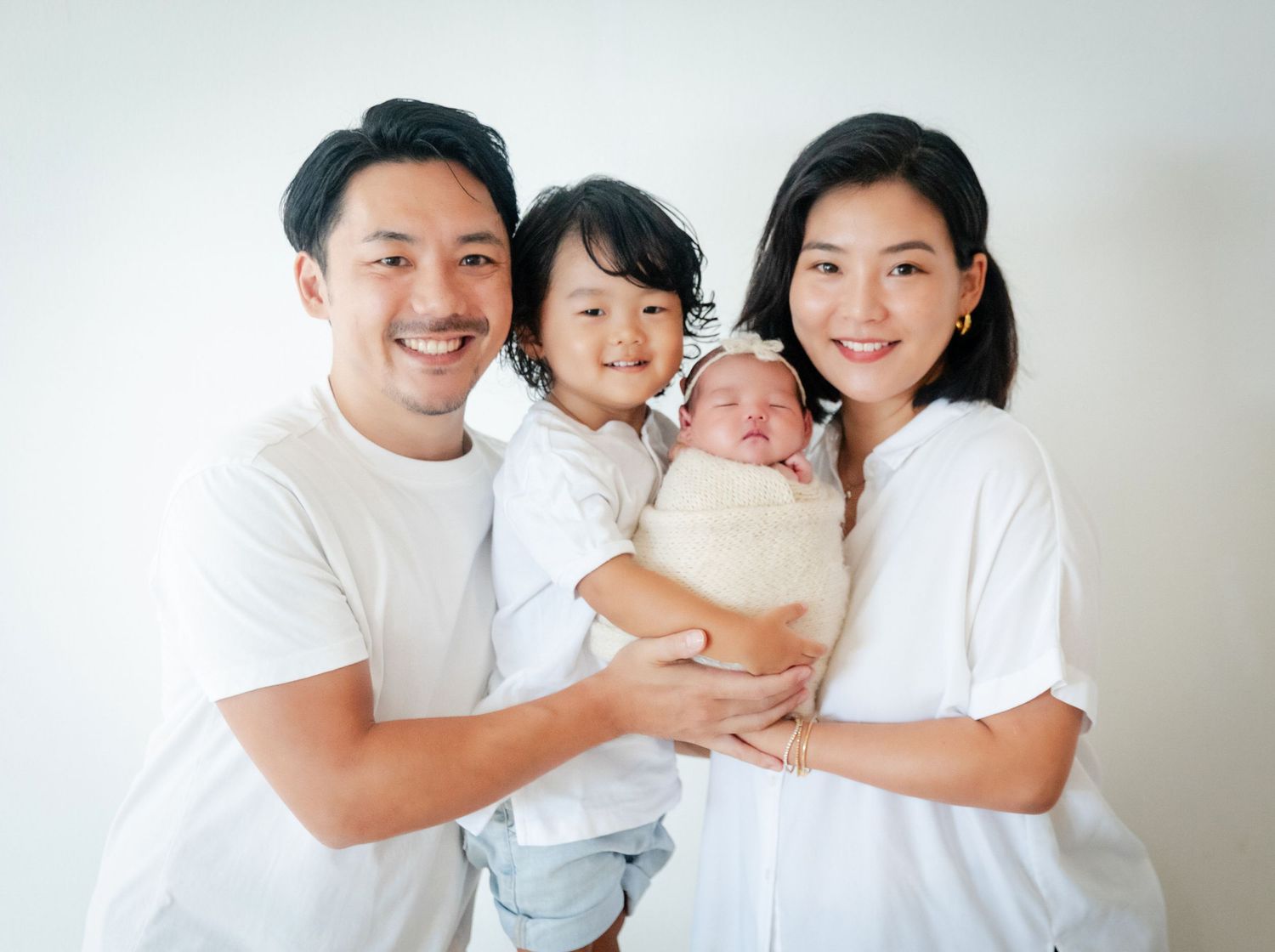 Happy Asian family of four posing together, parents holding toddler and swaddled newborn baby, wearing white shirts.