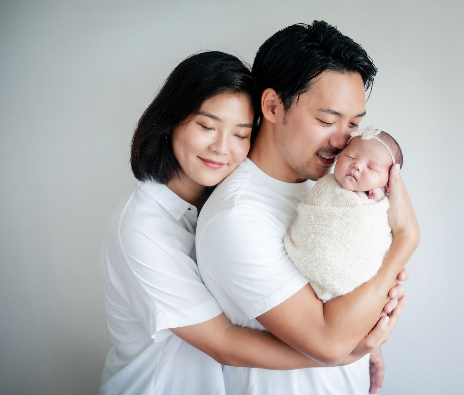 A young Asian couple in white shirts tenderly holds their swaddled newborn baby against a soft gray background.