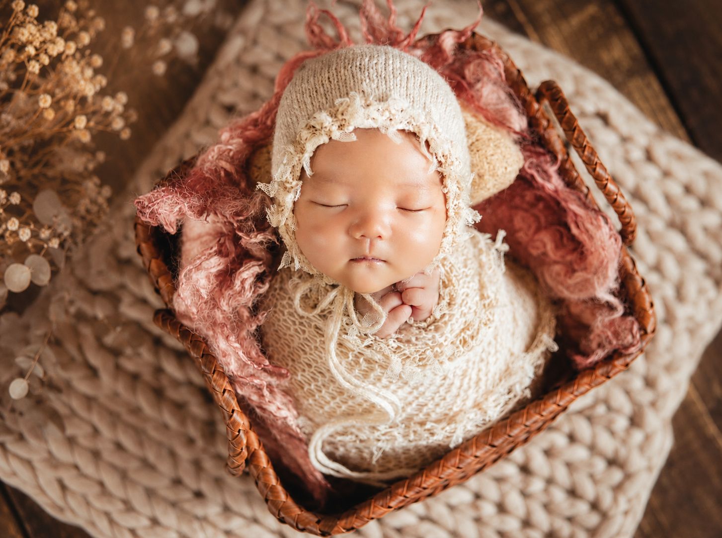 Sleeping newborn baby wrapped in cream knit swaddle wearing lace bonnet nestled in wicker basket with pink fabric.