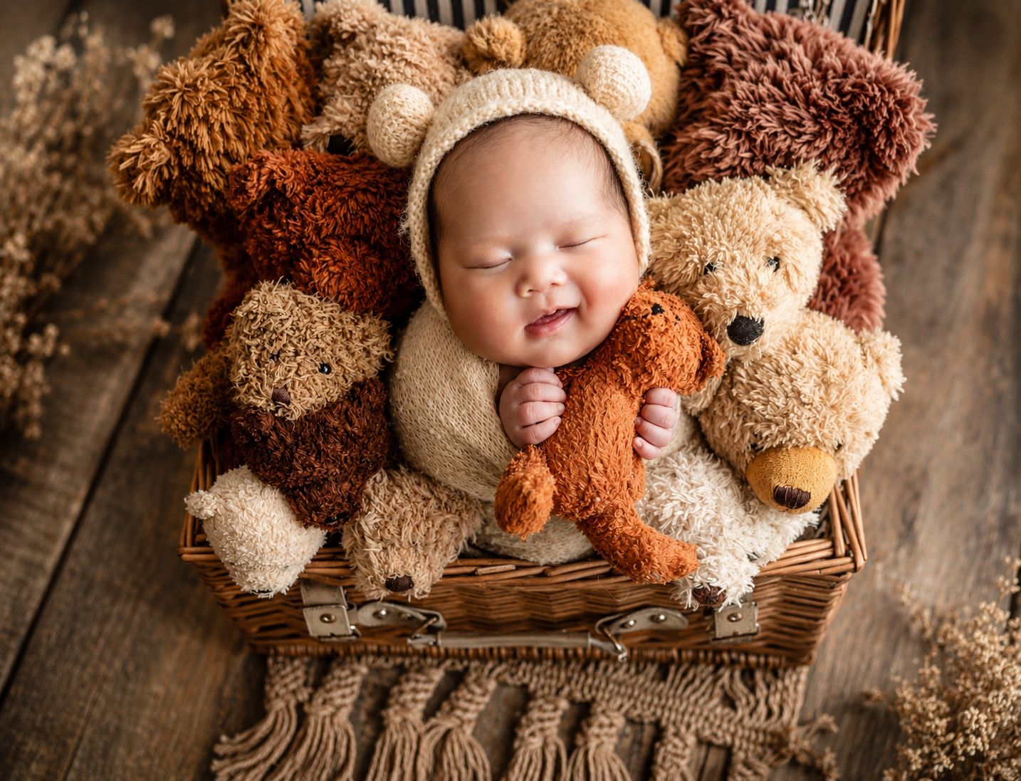 Smiling newborn baby in bear bonnet sleeping in wicker basket surrounded by teddy bears on rustic wood.