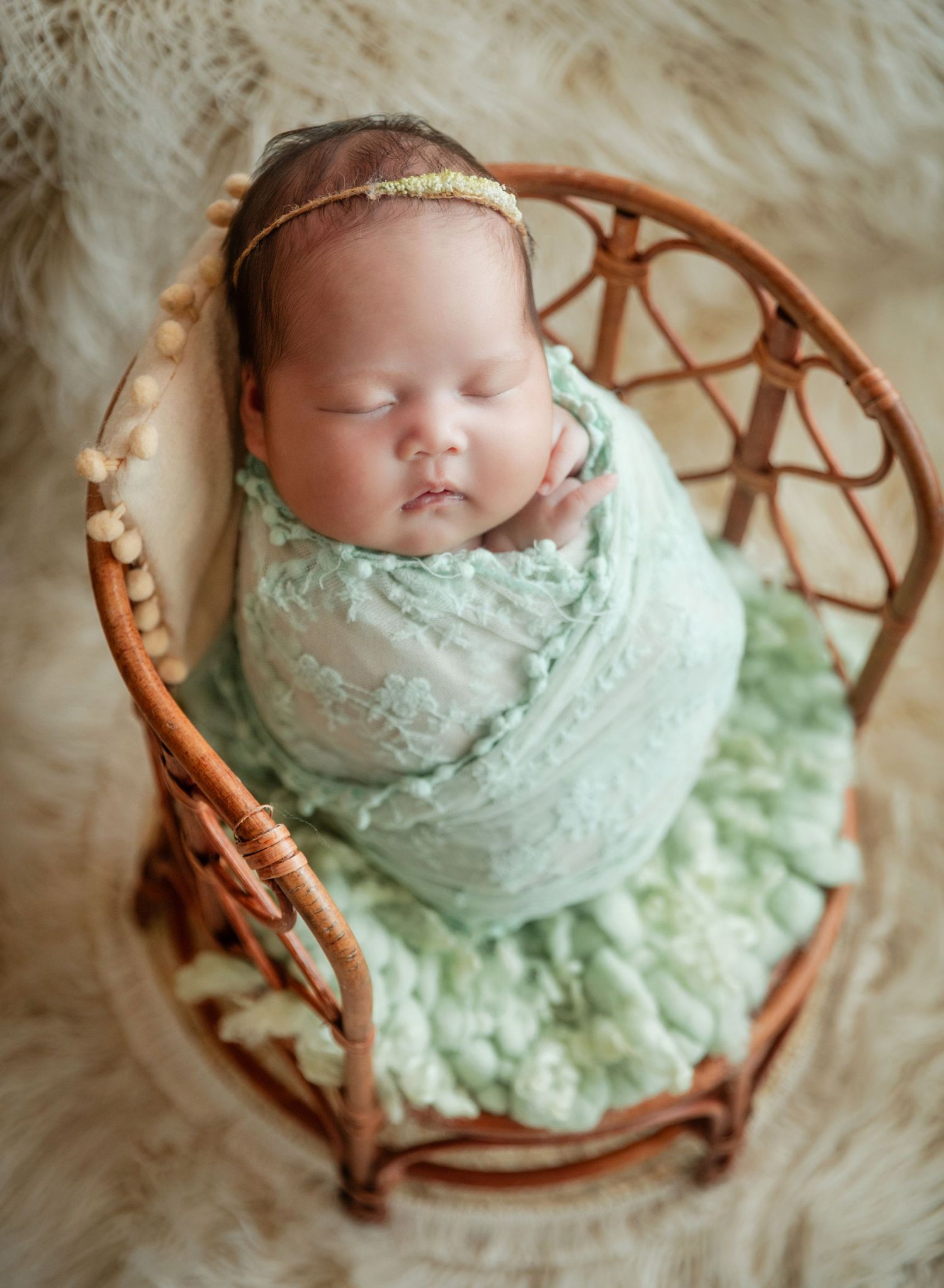 Newborn baby swaddled in mint green lace wrap, sleeping in a small wicker chair on a fluffy white rug.