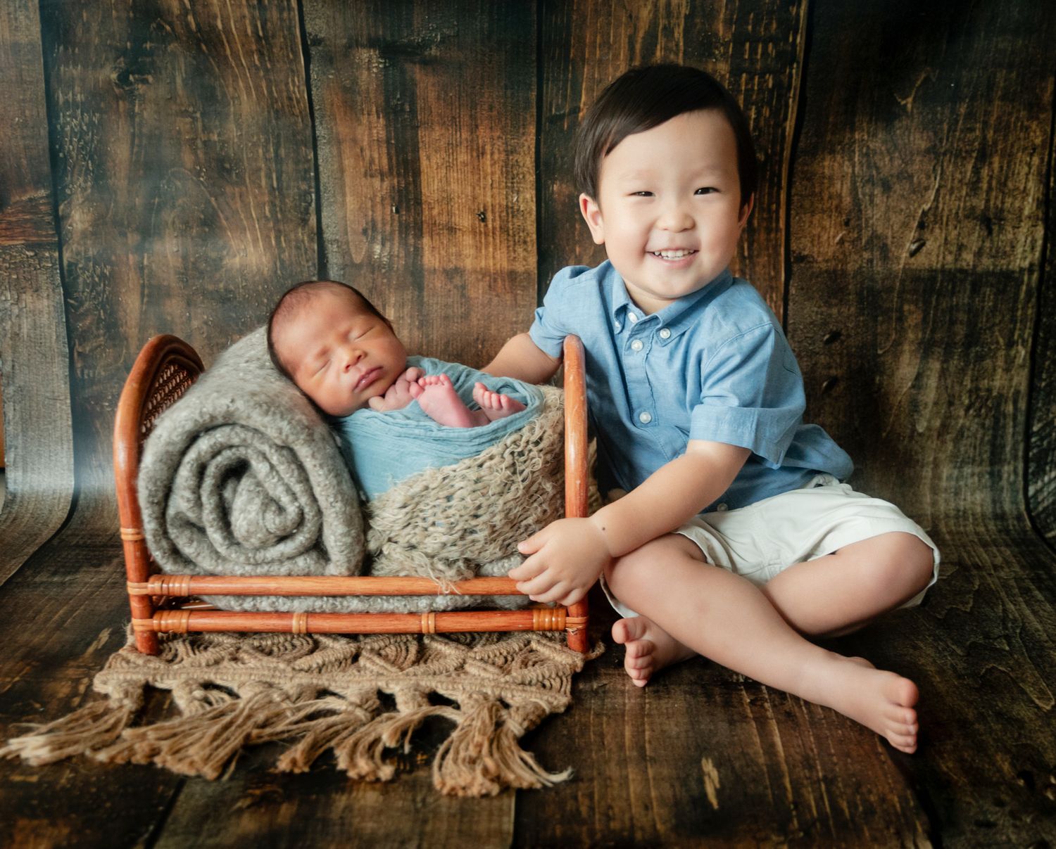 Smiling toddler boy poses beside sleeping newborn baby in tiny bed during professional photo session.