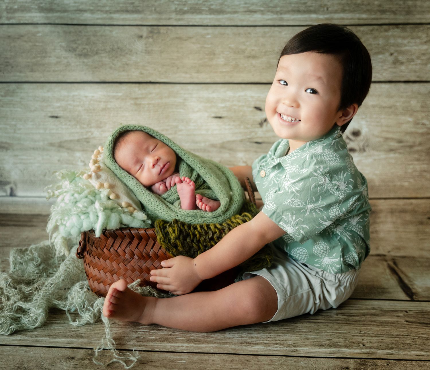 Smiling toddler boy holds a basket with a sleeping newborn wrapped in green knit, sibling photo session.