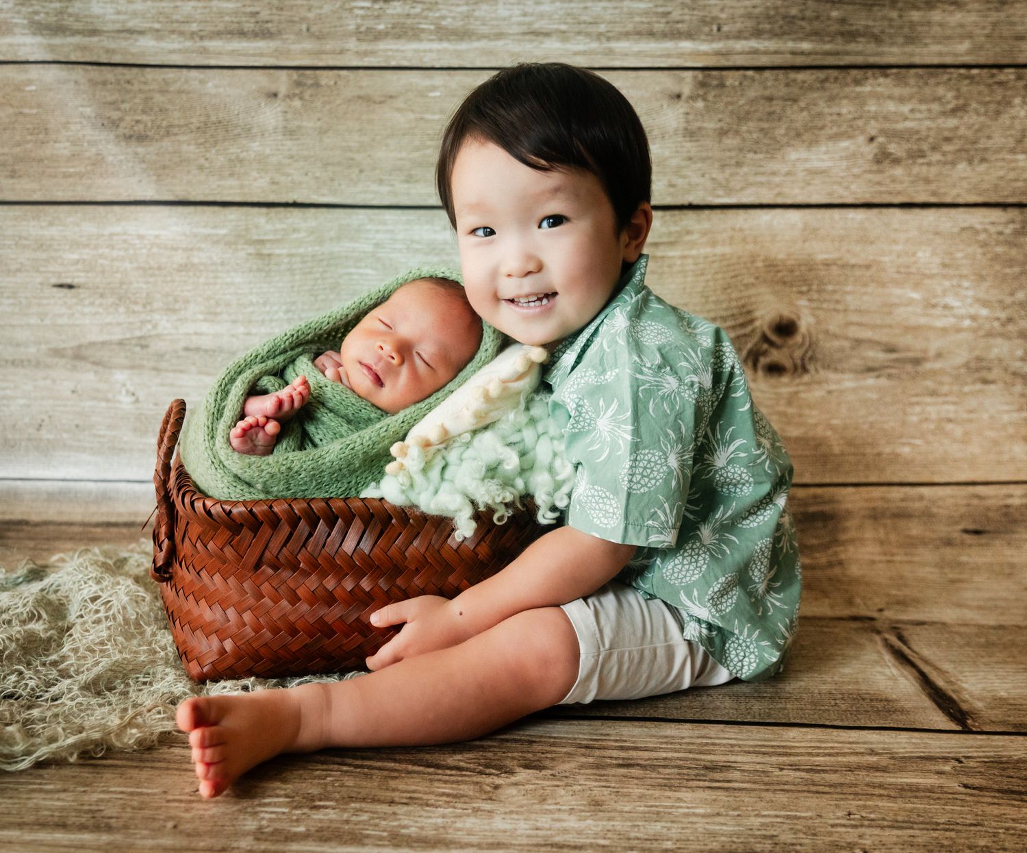 A smiling toddler boy in green shirt sits with a sleeping newborn baby wrapped in green in a wicker basket.