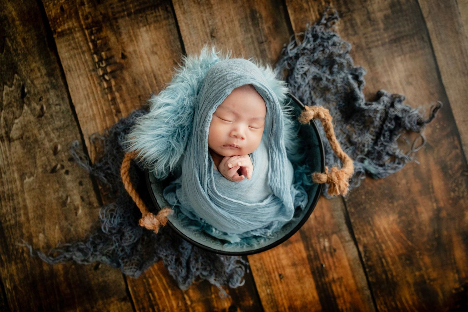 Newborn baby swaddled in blue wrap sleeping peacefully in a rustic wooden bucket with fluffy gray fur.