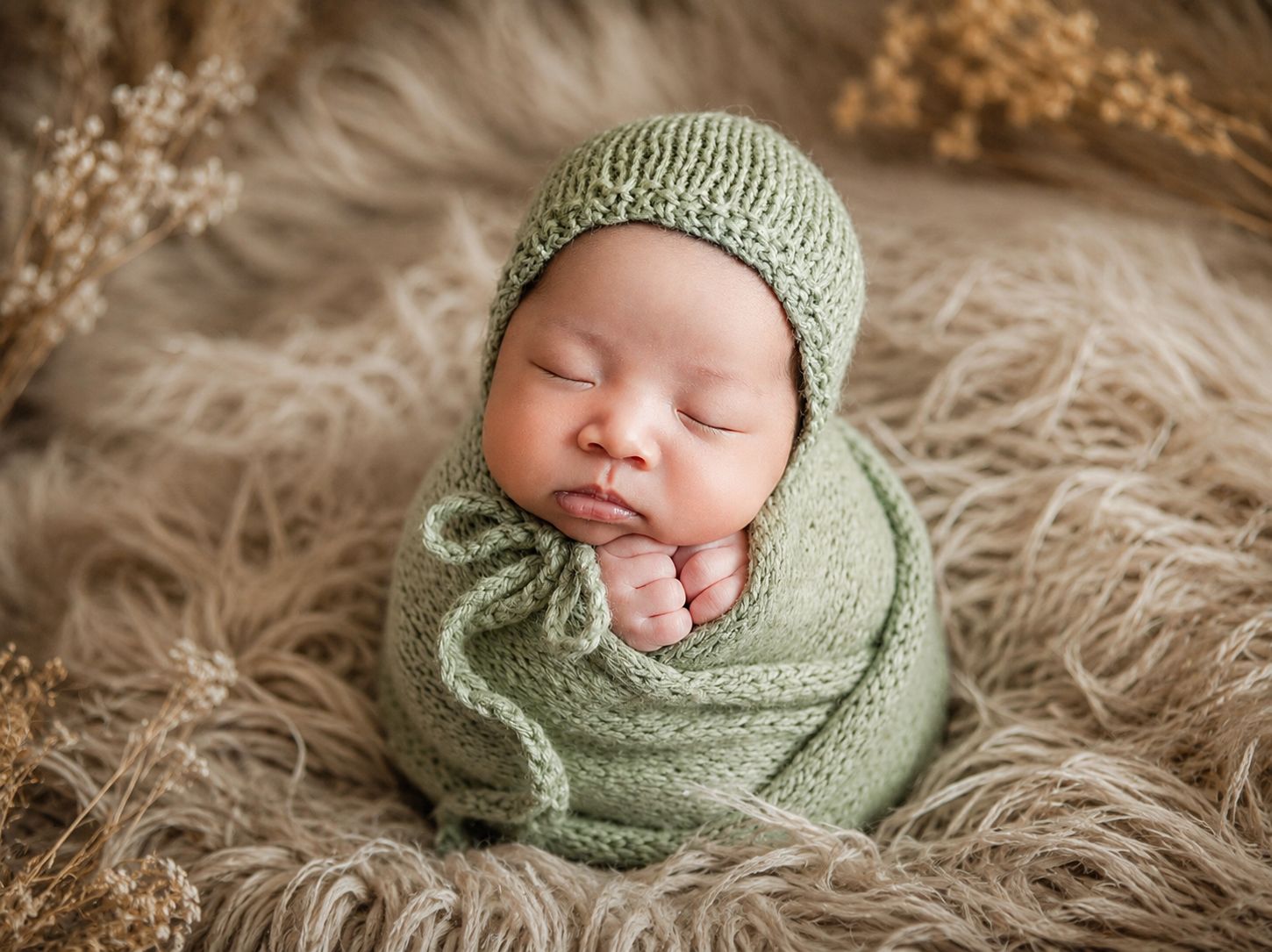 Sleeping newborn baby wrapped in green knit swaddle and matching bonnet hat on fluffy beige fur backdrop.