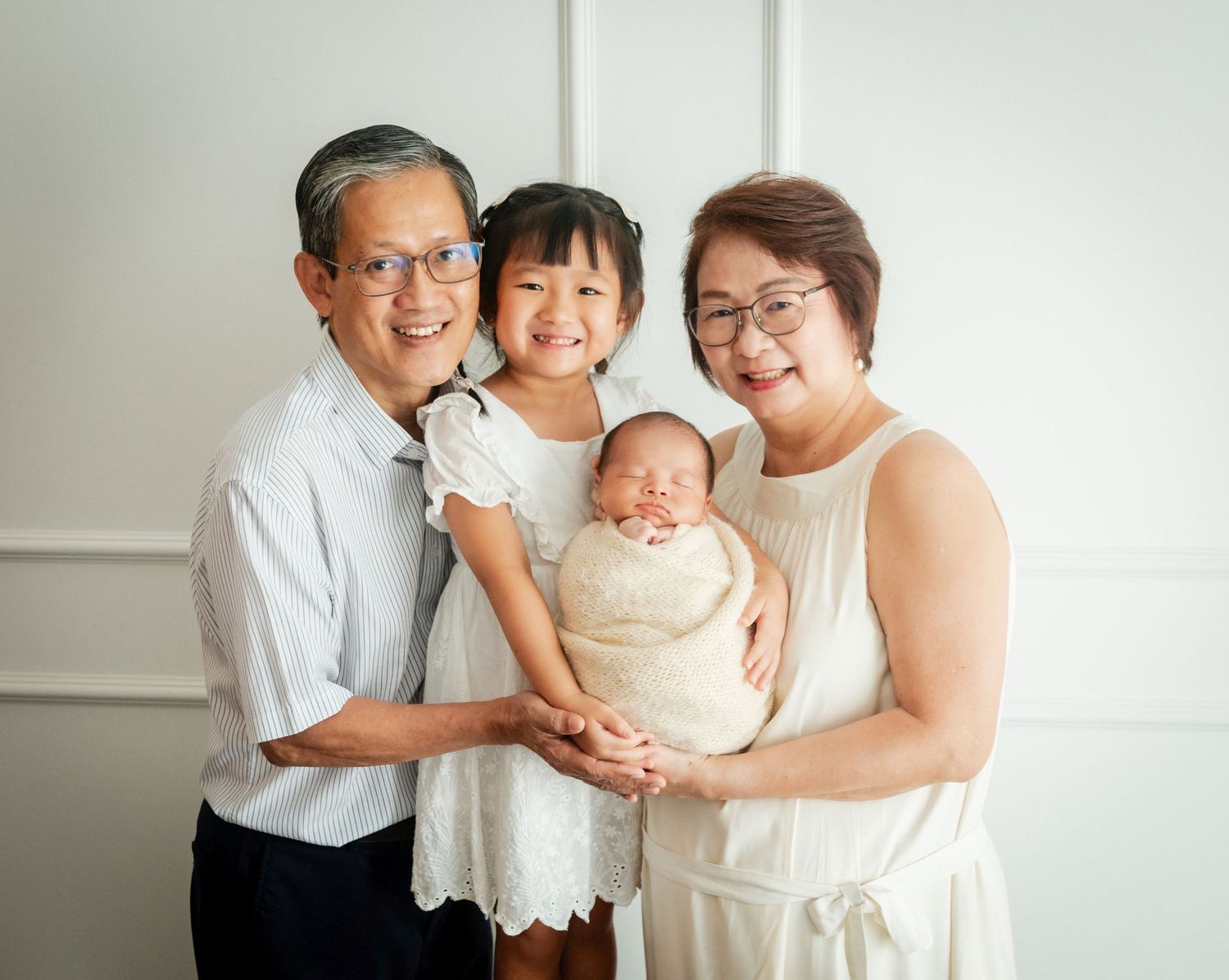 Grandparents smiling with granddaughter and newborn baby wrapped in cream swaddle during family portrait session.