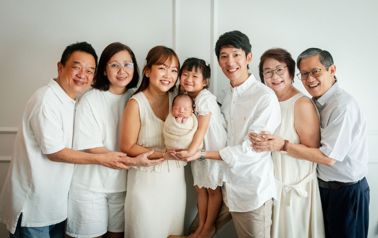 Multi-generational Asian family in white outfits smiling together, holding a newborn baby wrapped in white.