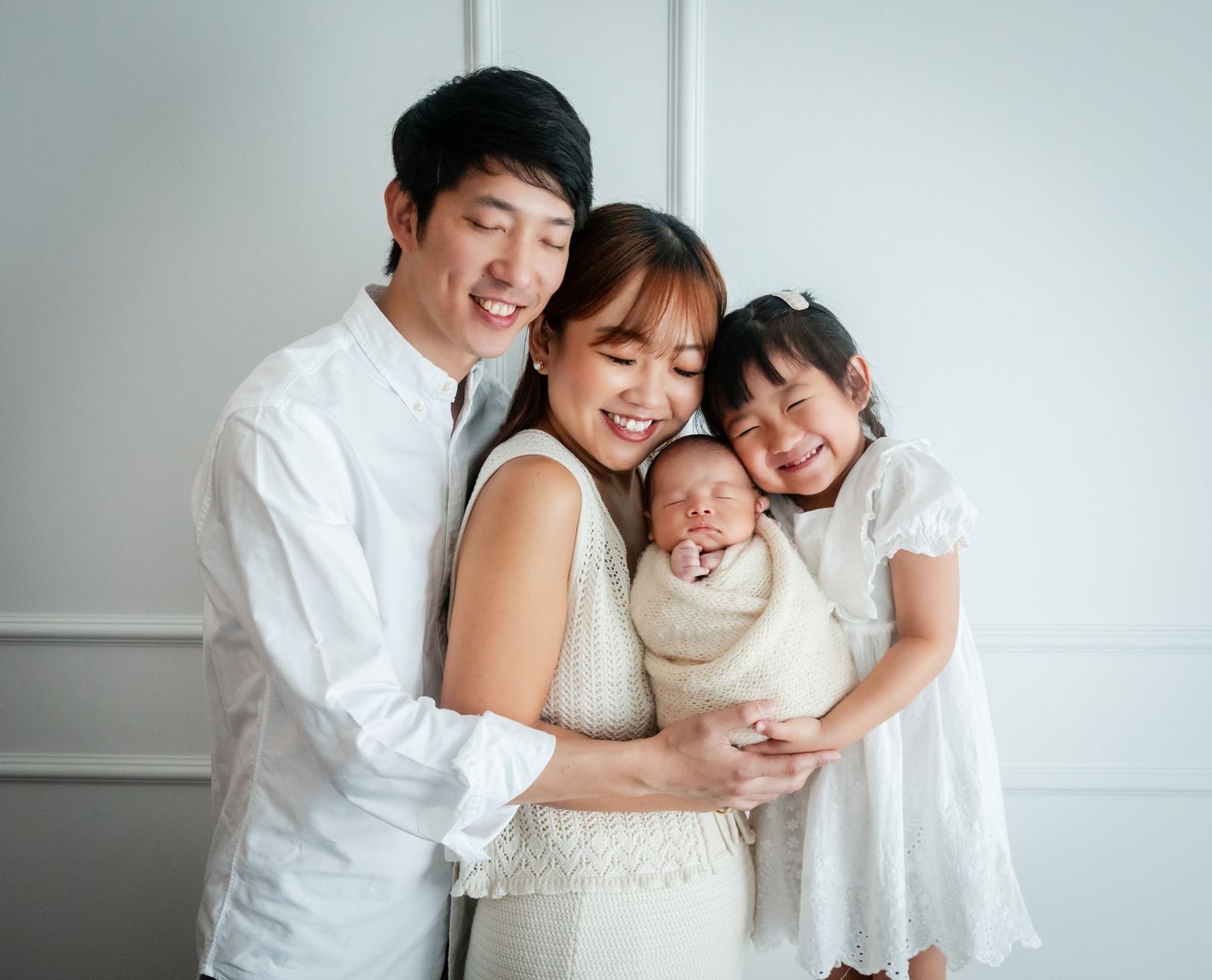 Happy Asian family of four posing together, parents holding newborn baby and toddler daughter, all wearing white outfits.