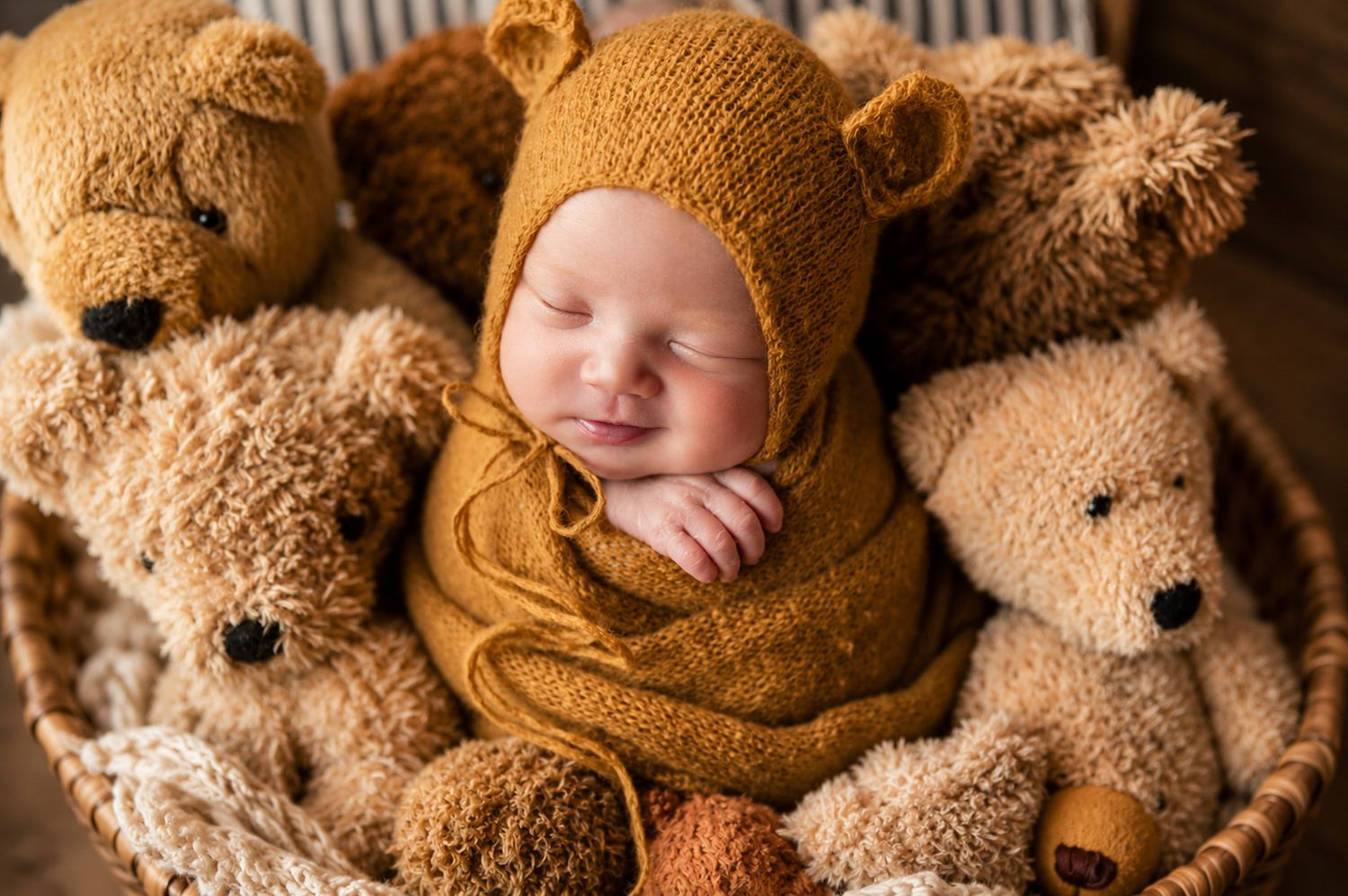 Newborn baby sleeping peacefully in a basket surrounded by brown teddy bears, wearing a cute knit bear bonnet.
