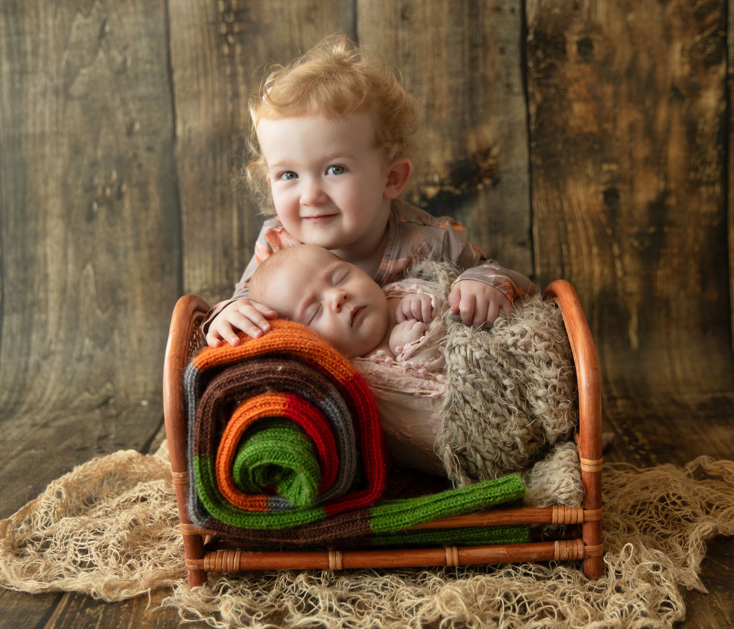 Toddler with curly hair smiles while holding a sleeping newborn in a small wooden bed with colorful rolled blankets.