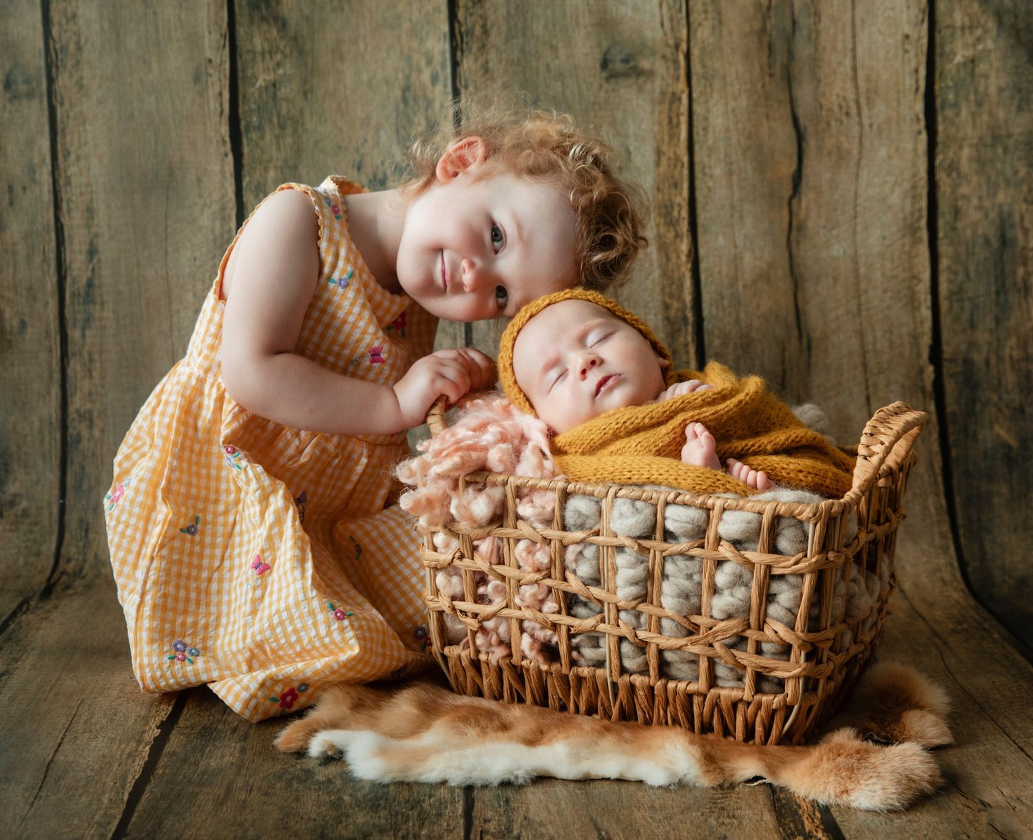 A smiling toddler girl in a yellow dress poses beside a sleeping newborn baby wrapped in a mustard blanket in a wicker basket.