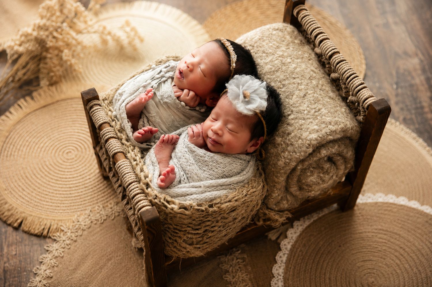 Two swaddled newborn twins sleeping peacefully together in a tiny rustic wooden bed with boho decor.