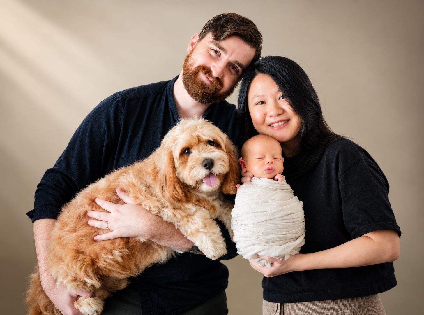 A smiling couple poses with a fluffy golden doodle dog and a swaddled newborn baby against a neutral background.