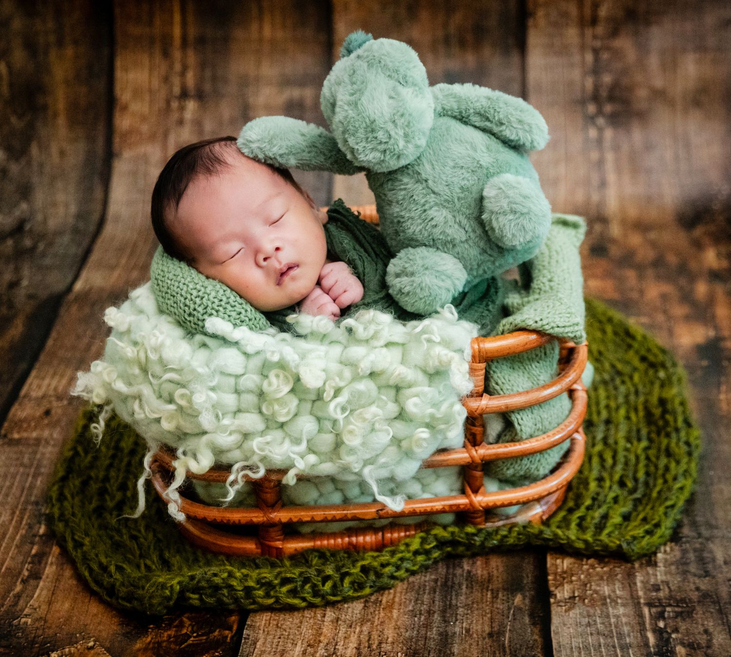 Sleeping newborn baby in a wicker basket with a green teddy bear and soft green blankets.
