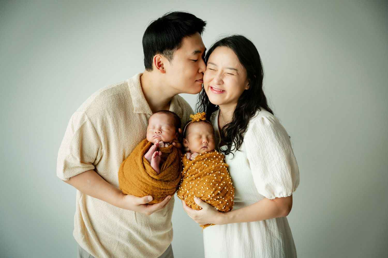 Asian couple holding newborn twins in a studio portrait, father kissing mother's cheek, babies wrapped in yellow swaddles.