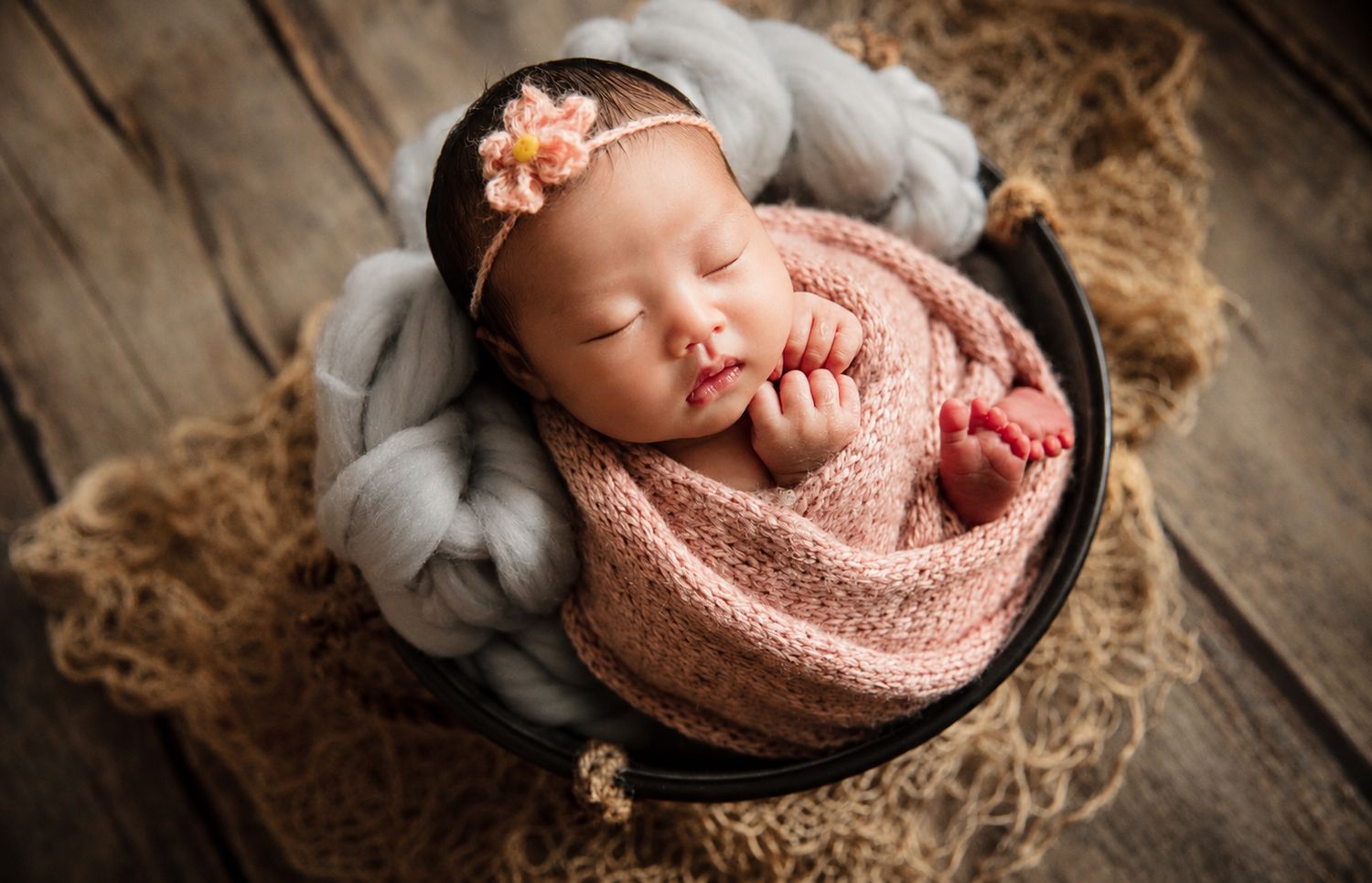 Peaceful newborn baby girl wrapped in pink knit blanket sleeping in a dark bucket with floral headband.