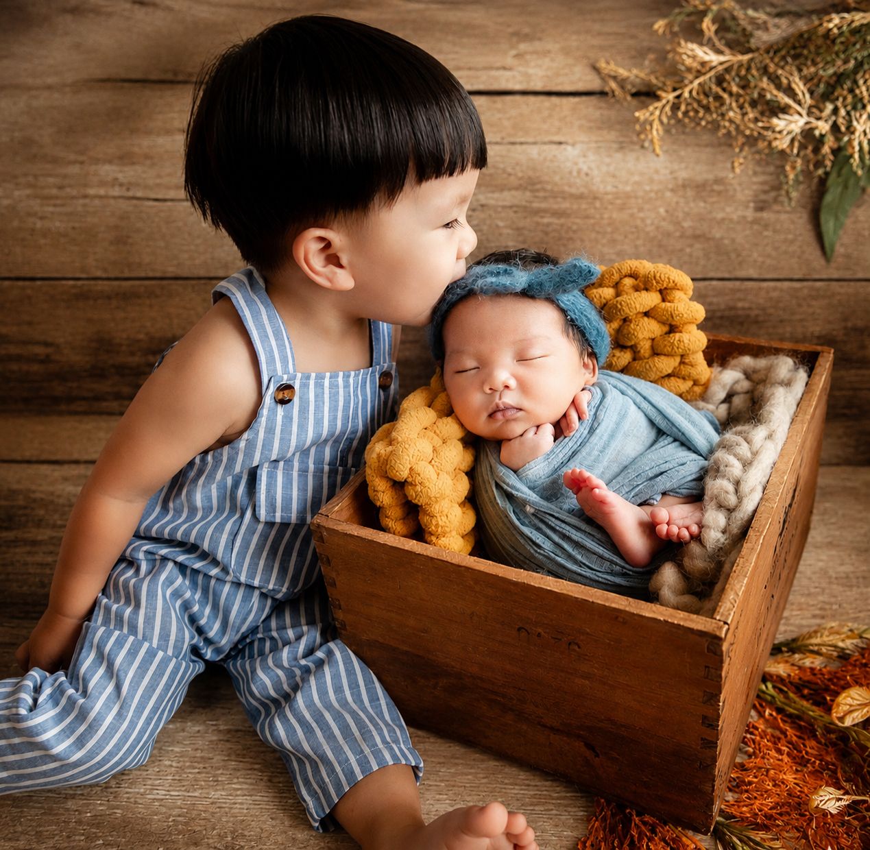 A young boy in striped overalls kisses a sleeping newborn wrapped in blue in a wooden crate.
