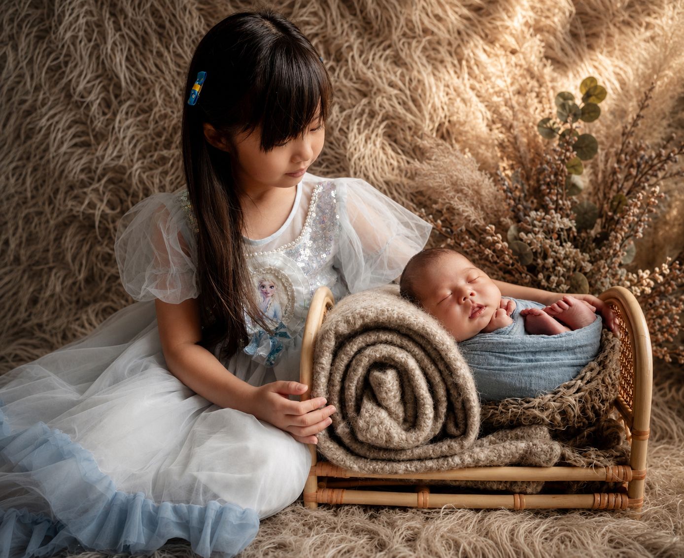A young girl in a blue dress gazes lovingly at a sleeping newborn in a rustic rattan bassinet with soft decor.
