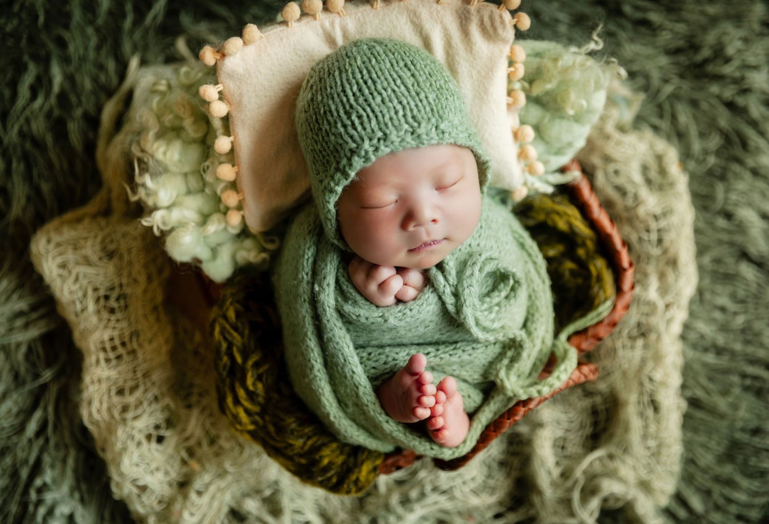 Newborn baby sleeping peacefully in a basket, wrapped in a green knit blanket and bonnet.