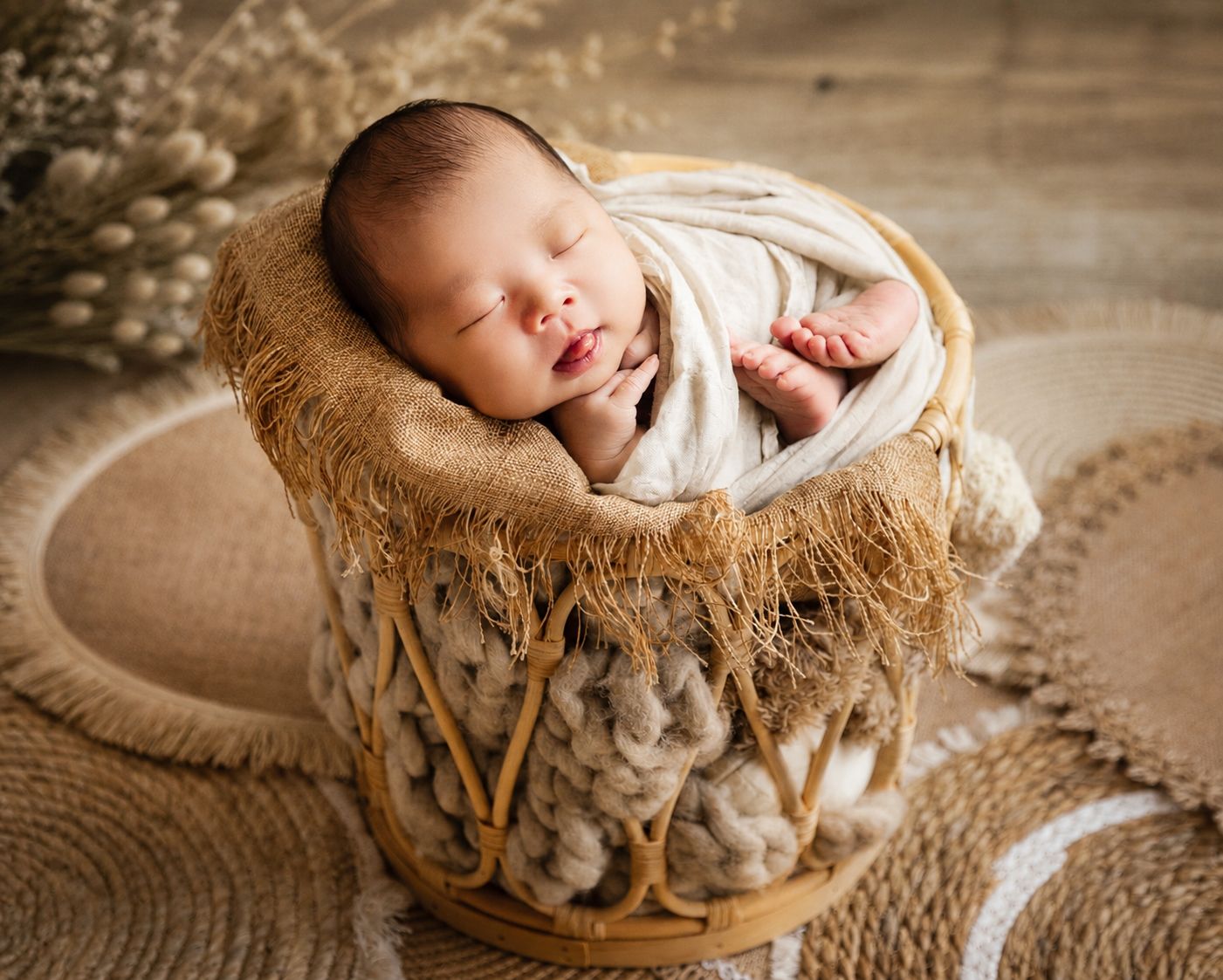 Smiling newborn baby wrapped in cream swaddle sleeping peacefully in a wicker basket with cozy knit blanket.