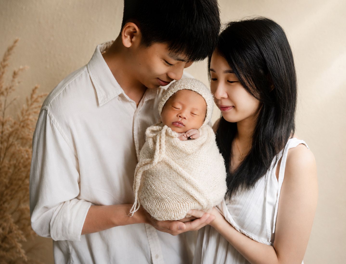 A young Asian couple tenderly holds their sleeping newborn baby wrapped in a cream knit swaddle.
