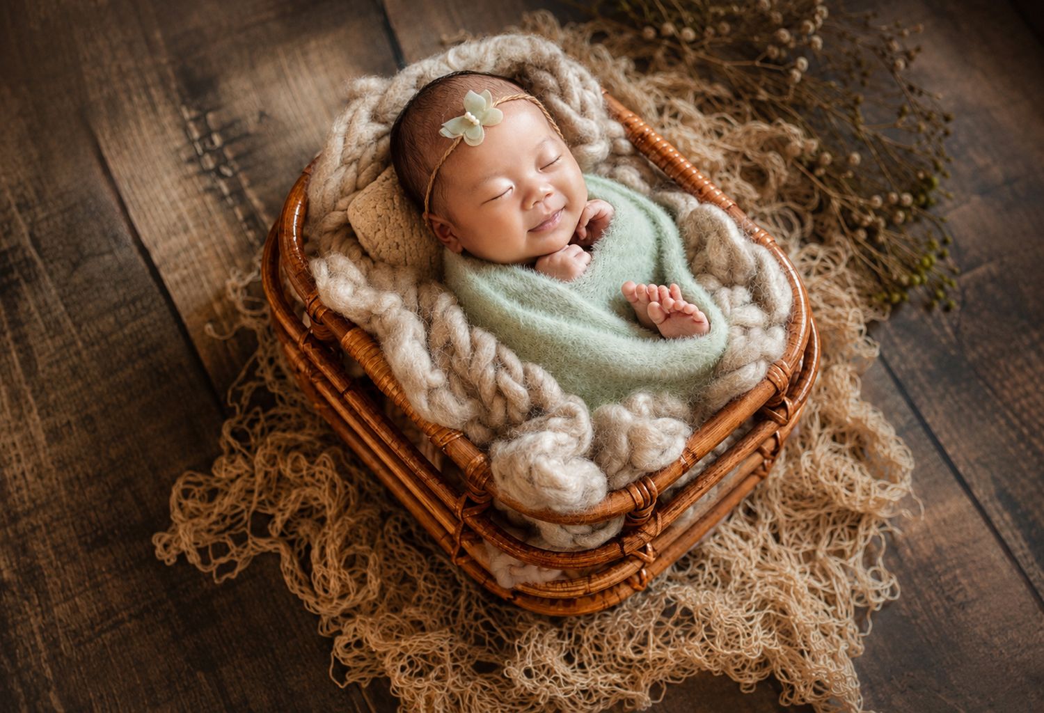 Smiling newborn baby sleeping in a wicker basket with chunky knit wrap and green bow headband.