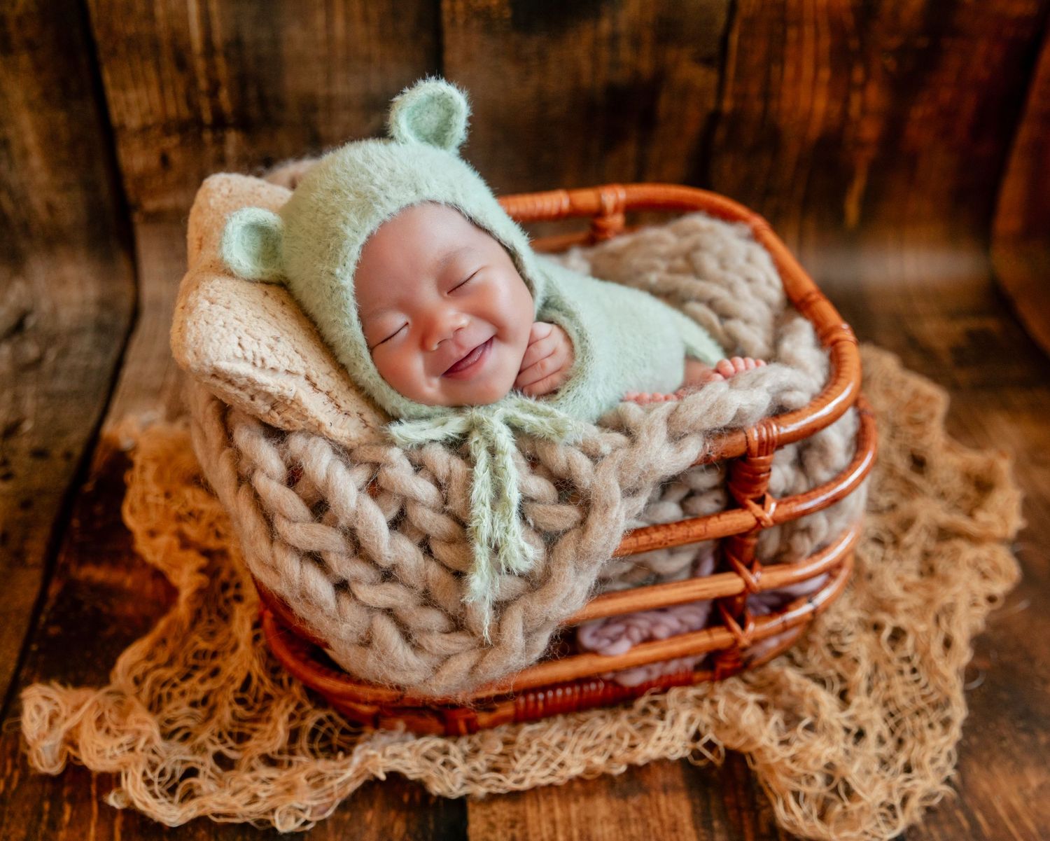 Smiling newborn baby in green bear bonnet sleeping in wicker basket with chunky knit blanket.