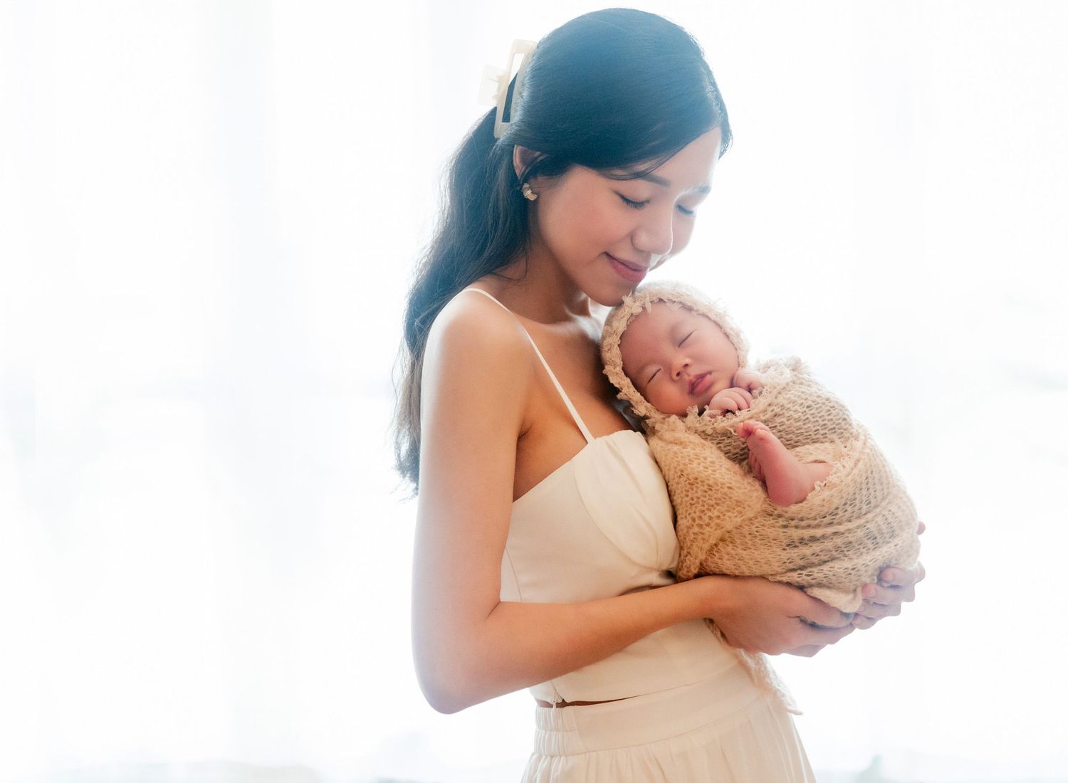 A young mother tenderly holds her sleeping newborn baby wrapped in a knit blanket near a bright window.