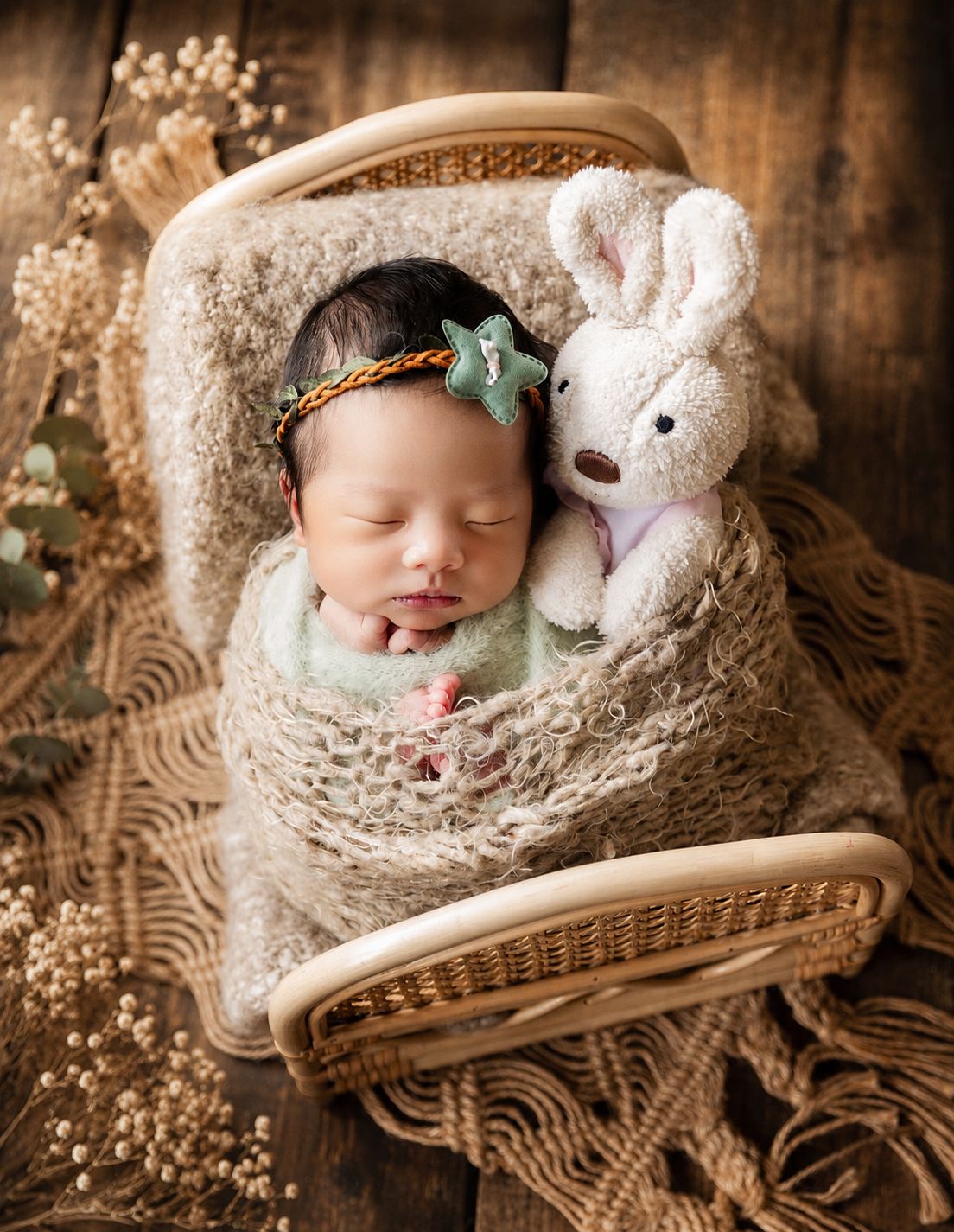 Newborn baby sleeping in wicker basket wrapped in beige knit blanket with star headband and white bunny stuffed animal.