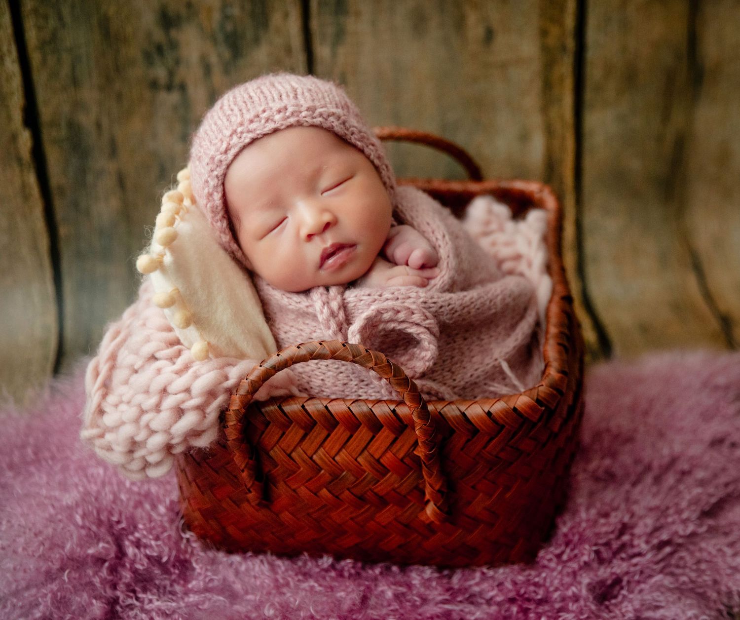 Newborn baby sleeping peacefully in a woven basket, wrapped in a soft pink knit blanket and matching hat.