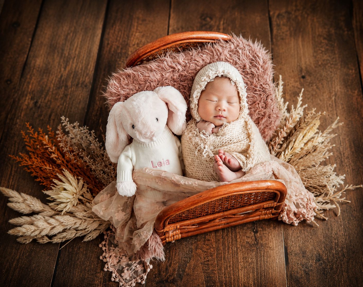 Sleeping newborn baby in cream wrap with bonnet cuddles white bunny toy in wicker basket with dried florals.