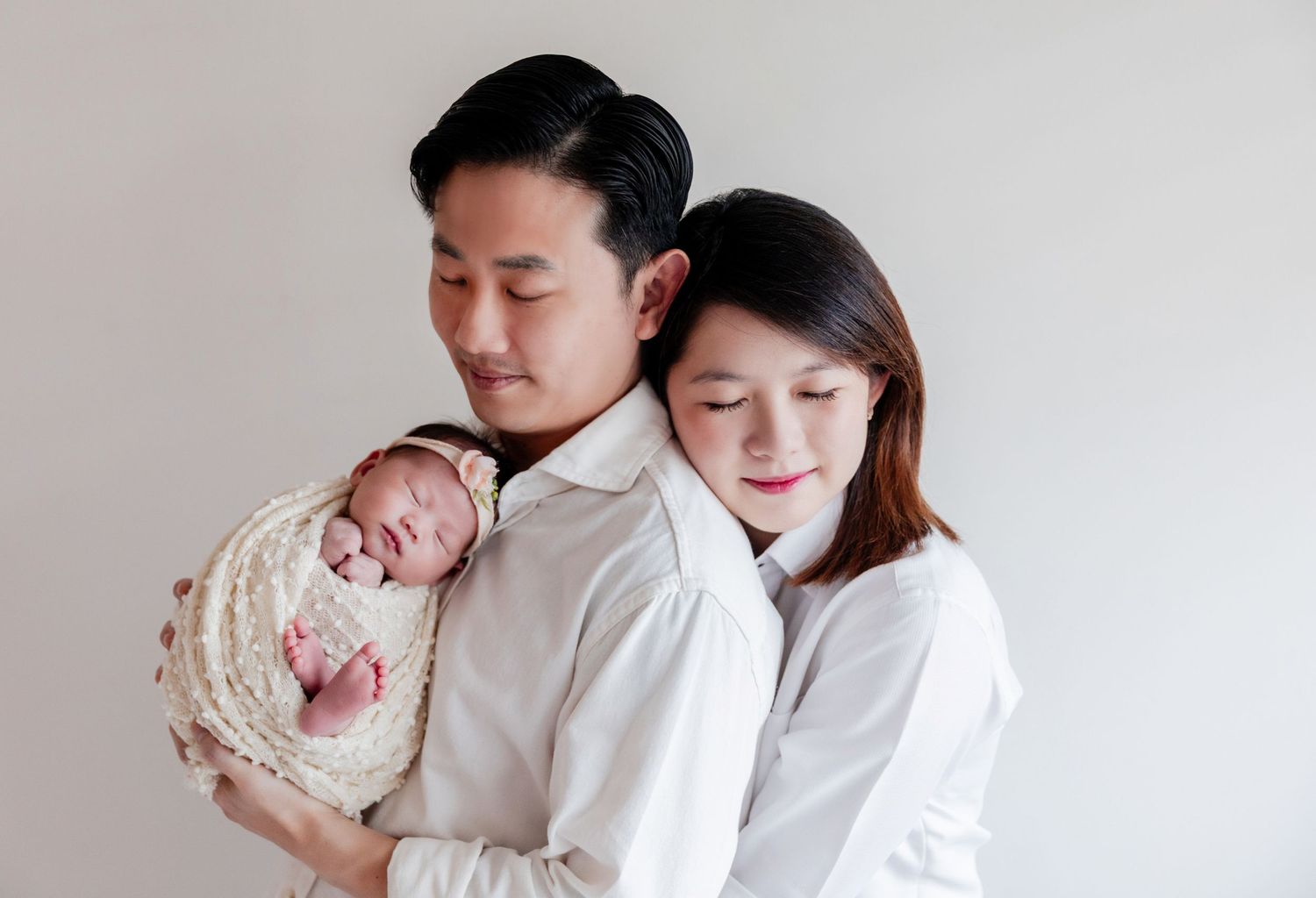A couple tenderly holds their sleeping newborn baby wrapped in a white knit blanket against a neutral background.