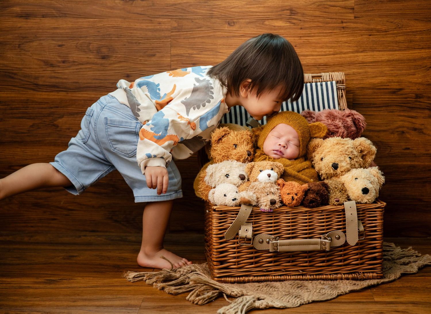 Toddler in dinosaur pajamas kisses sleeping newborn nestled among teddy bears in a wicker basket on wood floor.