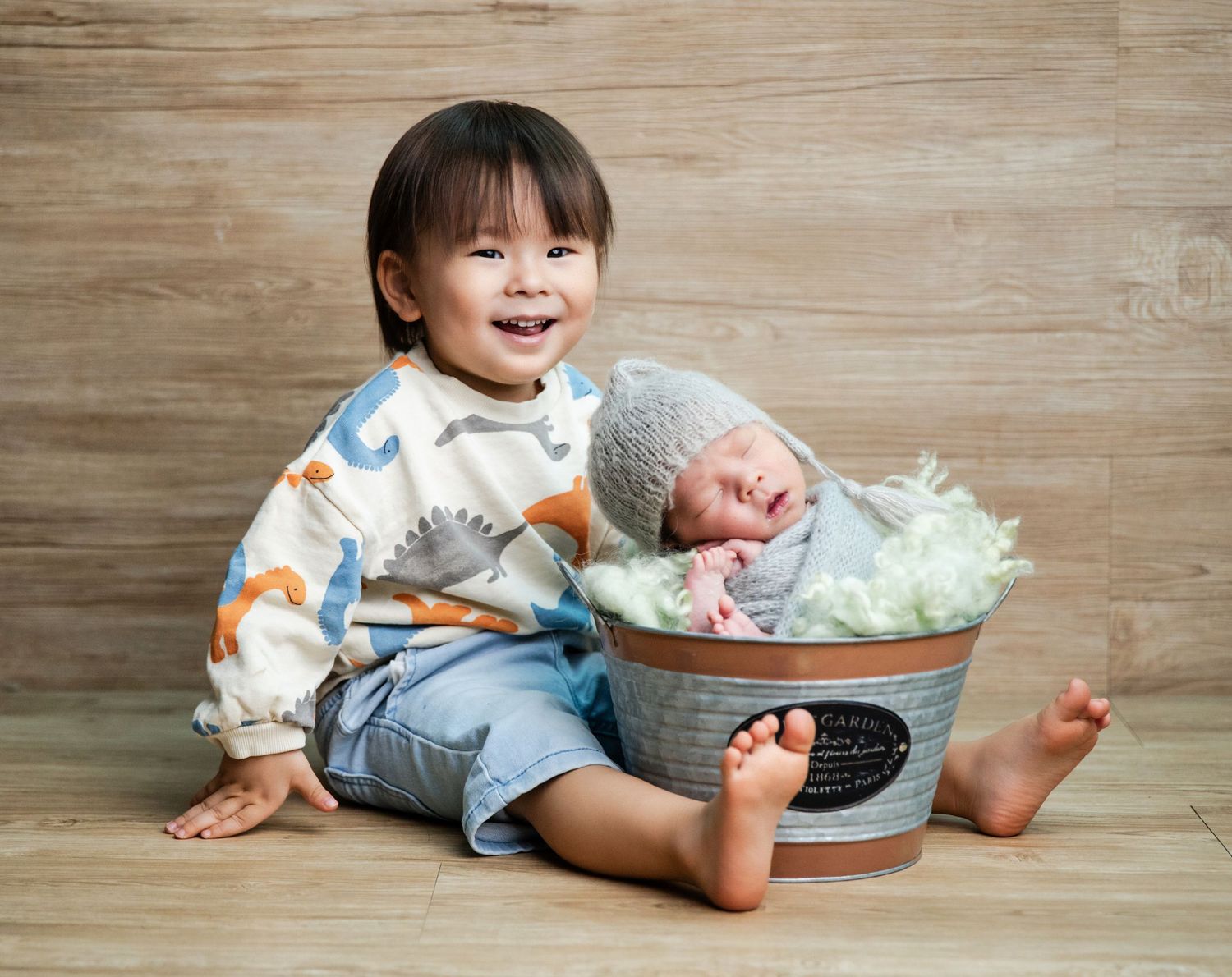 Smiling toddler sitting beside a newborn baby sleeping in a metal bucket with green filler for a sibling photo.
