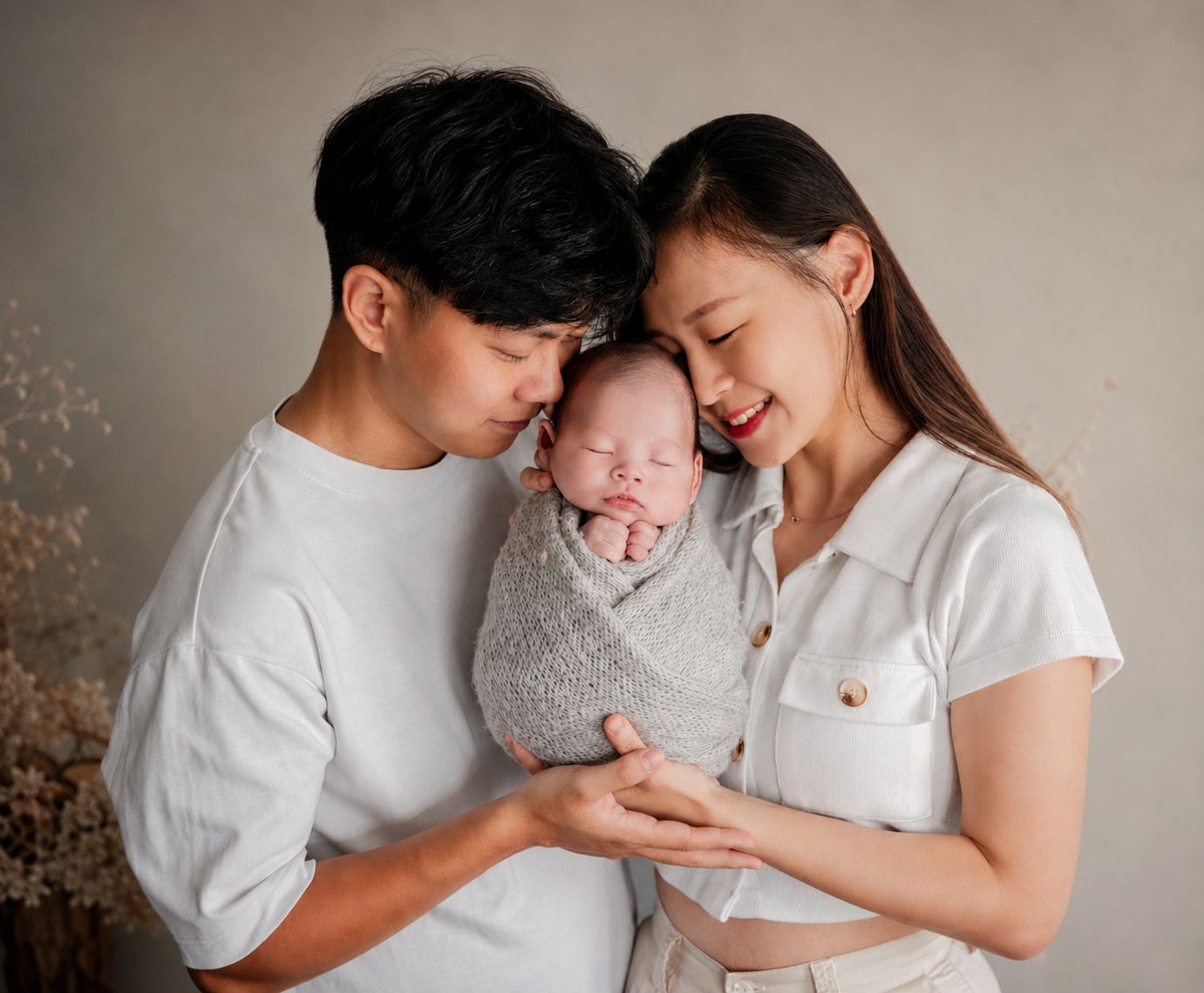 A couple in white outfits tenderly holds their swaddled newborn baby during a professional studio photo session.