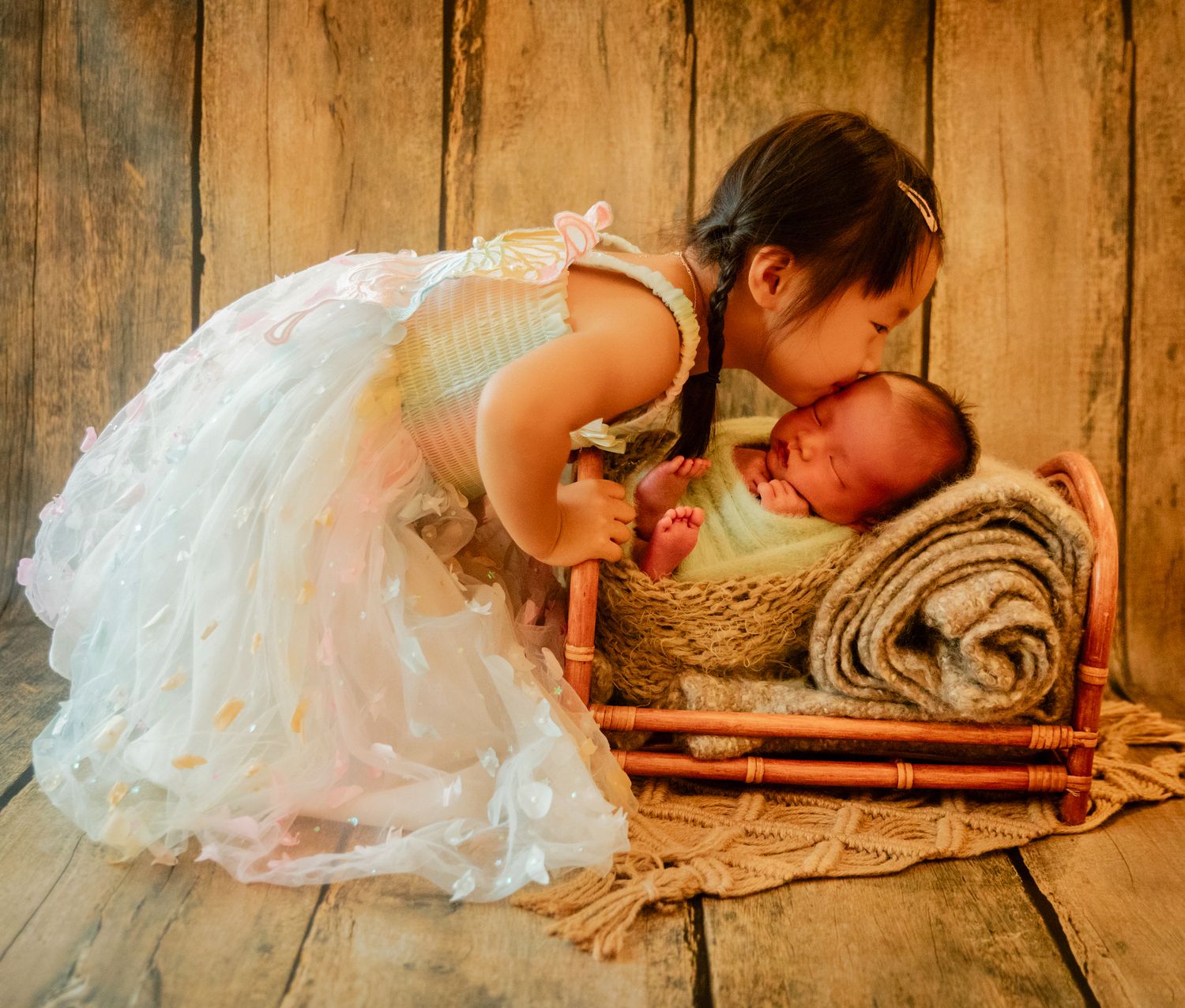 A young girl in a white dress gently kisses a sleeping newborn baby in a rustic wicker basket on a wooden floor.