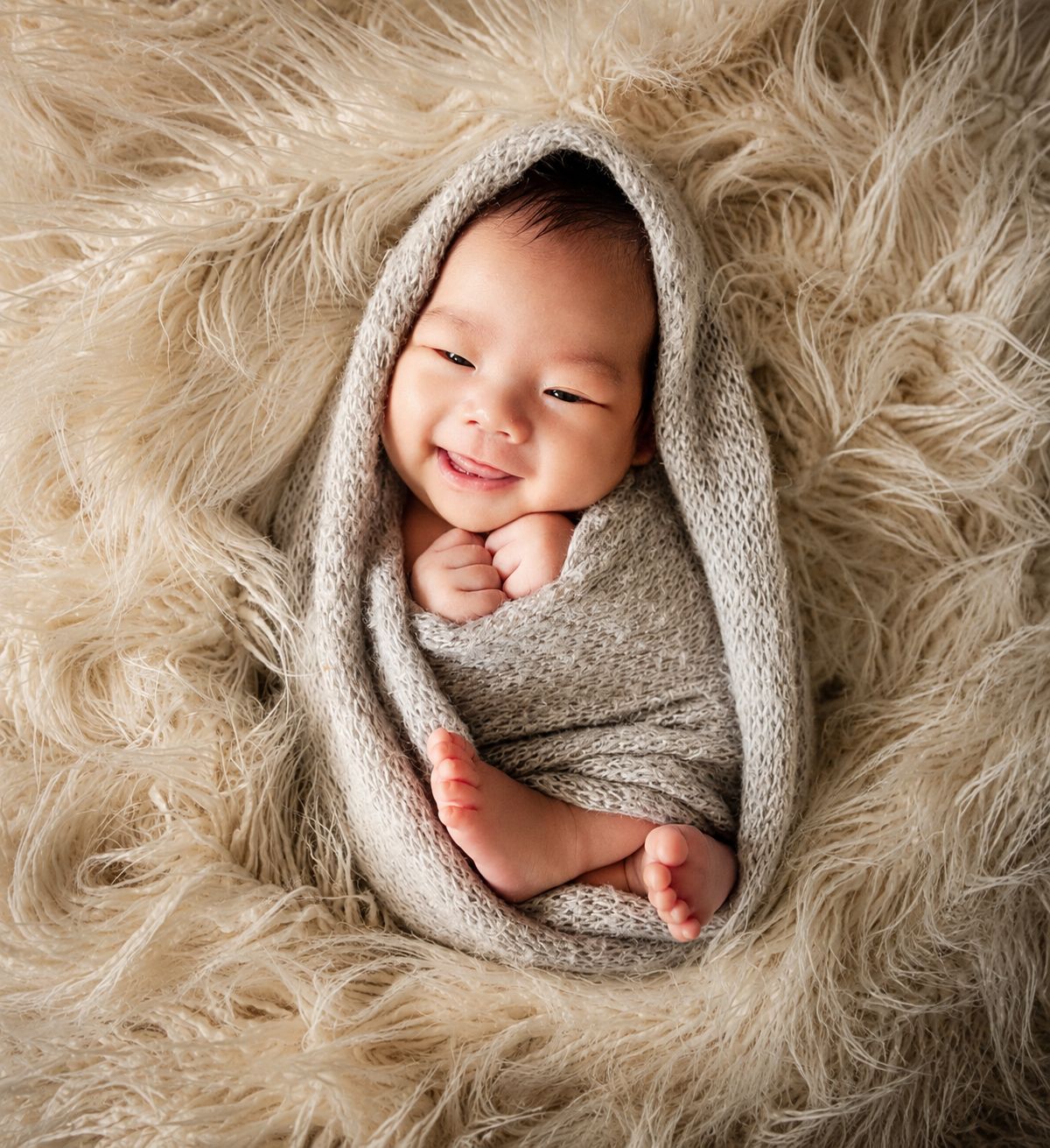 Smiling newborn baby swaddled in a beige knit wrap, nestled in a fluffy cream fur blanket.
