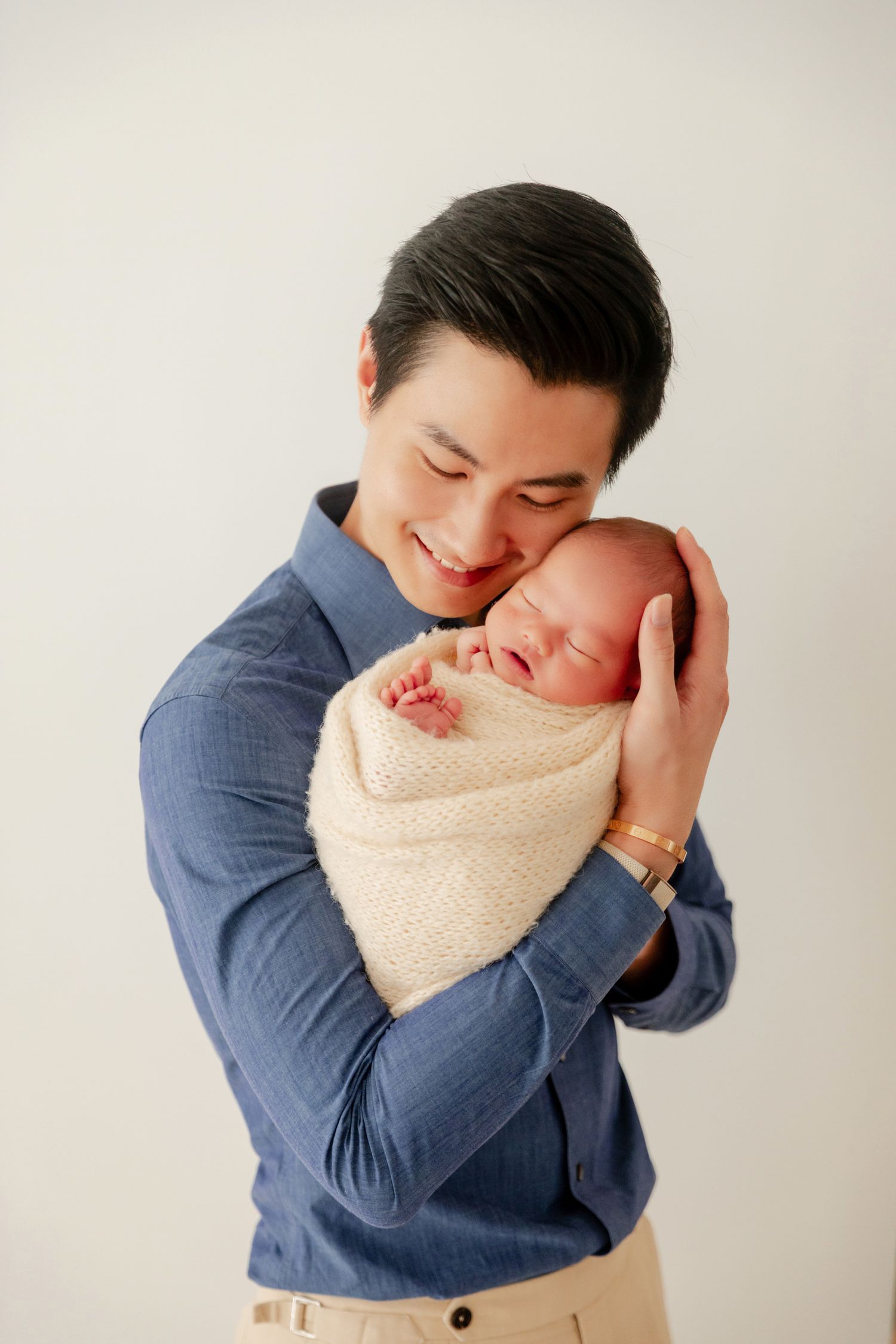 Smiling Asian man in blue shirt tenderly holds sleeping newborn baby wrapped in cream knit blanket.