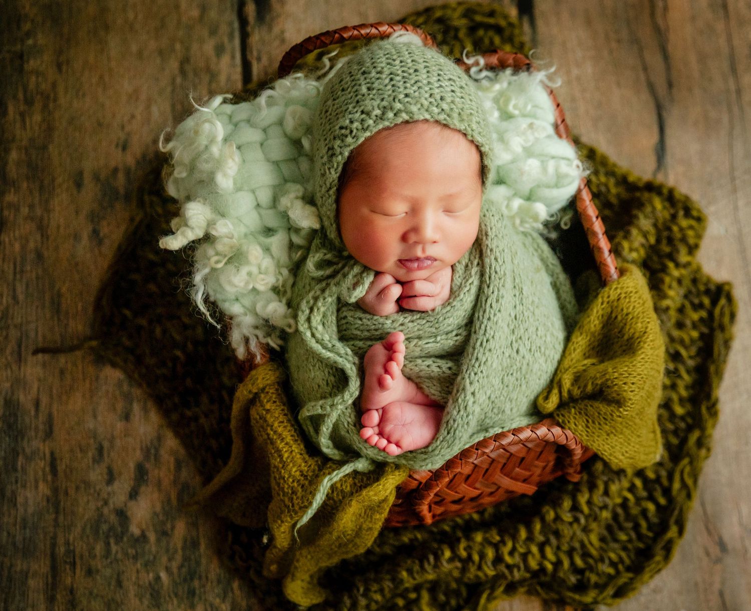 Newborn baby sleeping peacefully in a basket, wrapped in a green knit swaddle and matching bonnet hat.