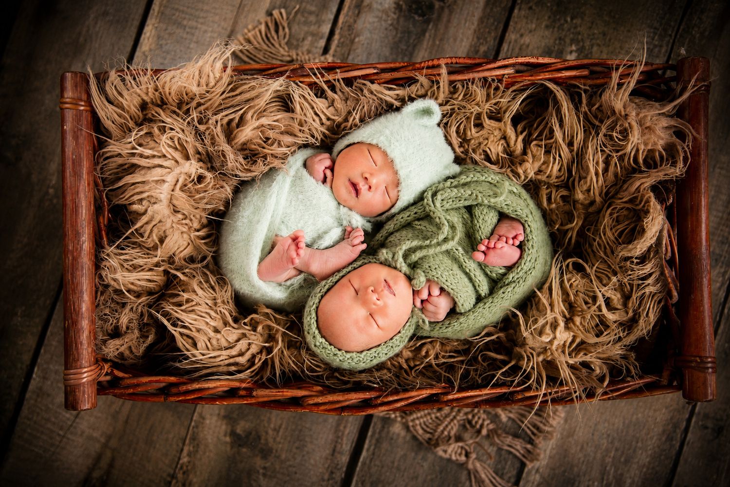 Two sleeping newborn twins wrapped in green knit swaddles rest in a rustic wicker basket lined with straw.